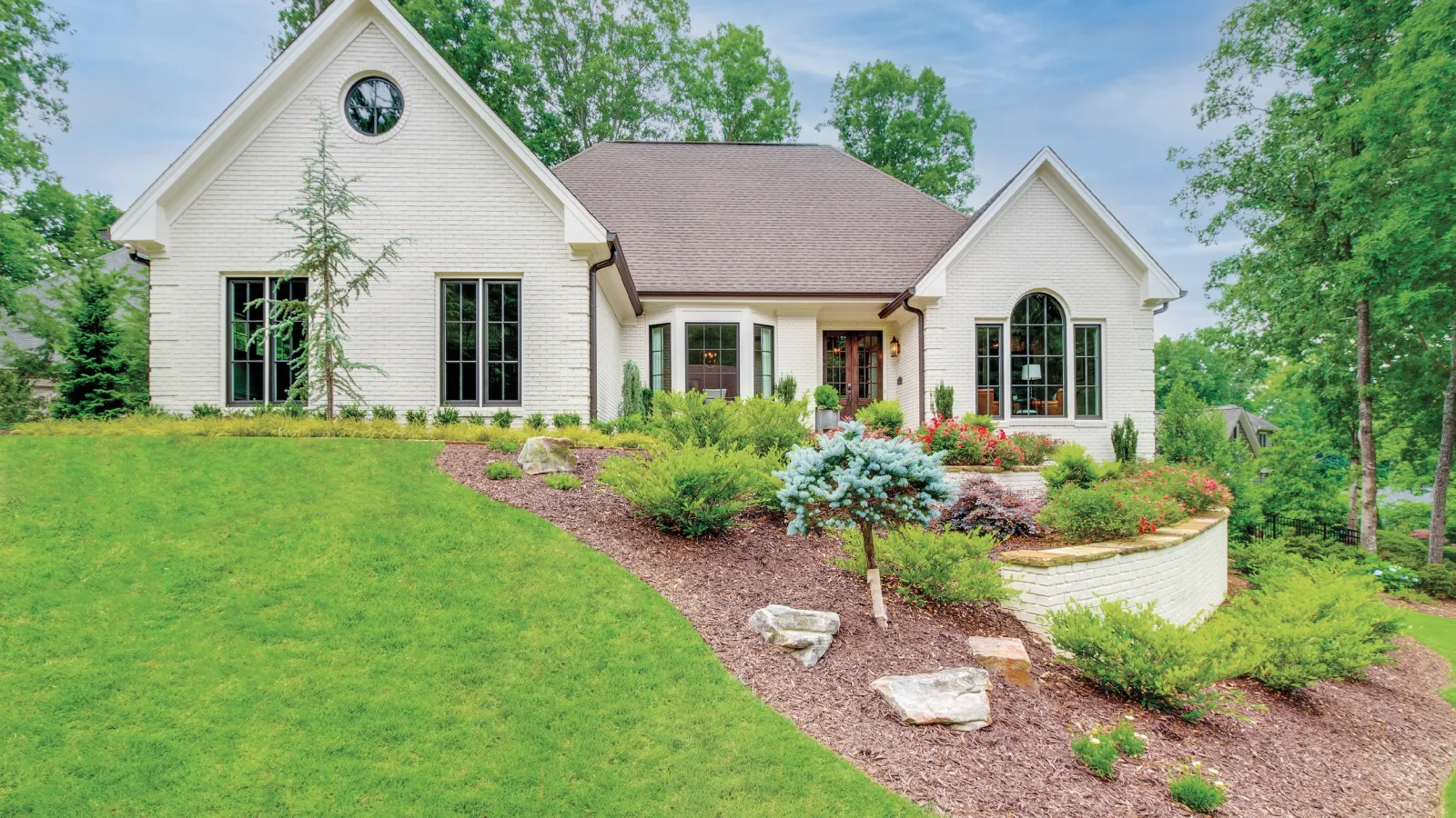White brick house with large windows surrounded by lush green lawn and landscaped garden beds