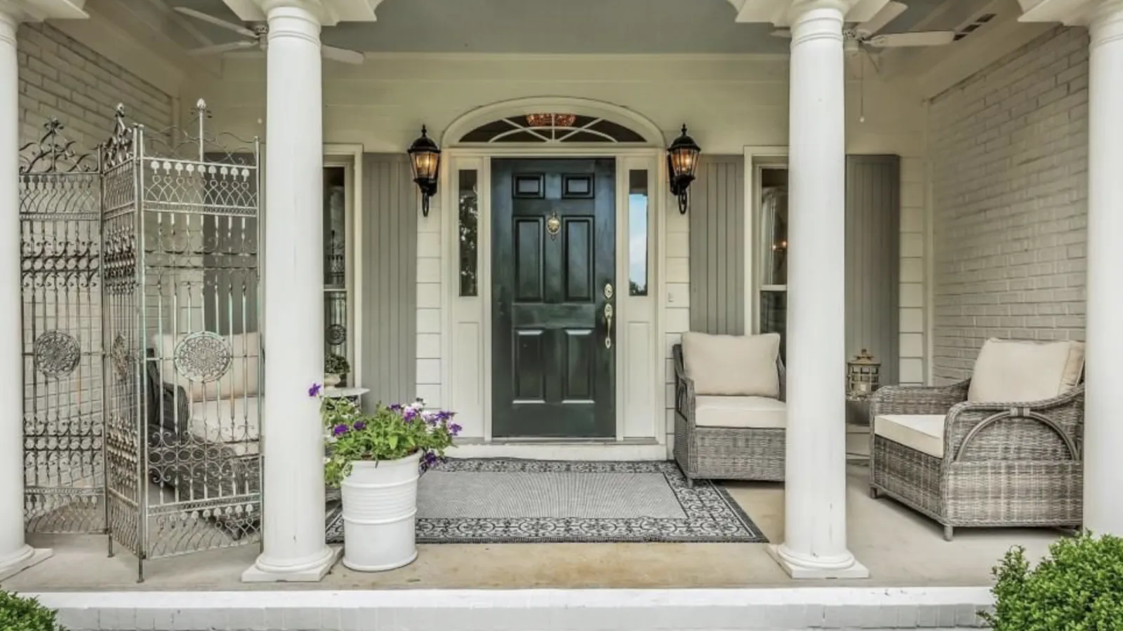 Elegant front porch with white columns, black door, gray wicker chairs, metal screen, and potted flowers.