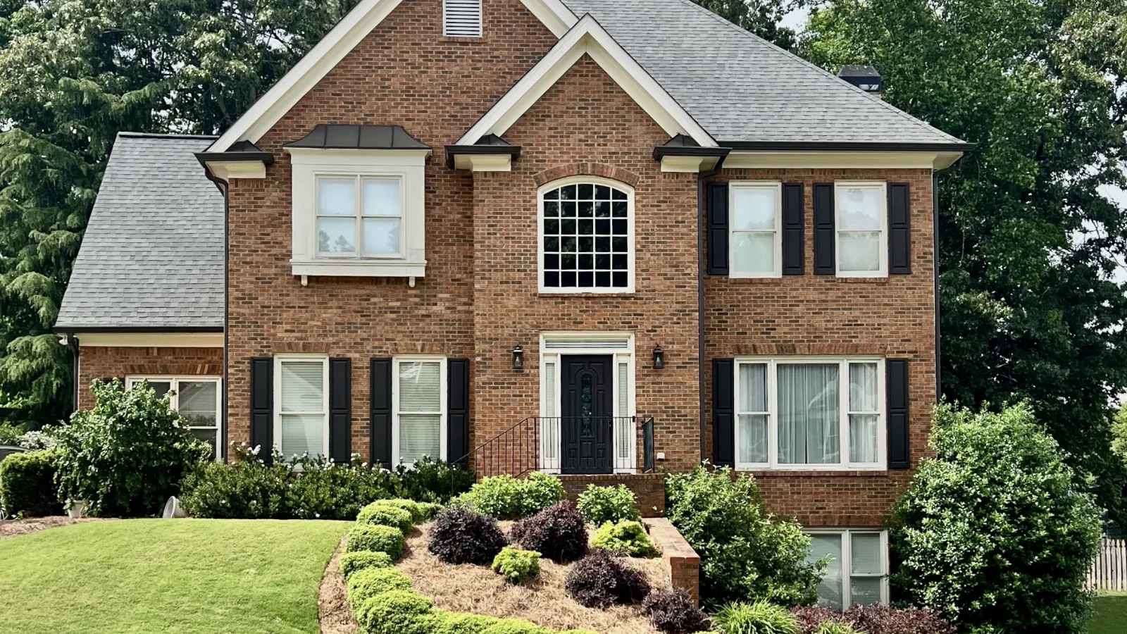 Two-story brick house with black shutters, a black door, gray roof, and manicured lawn with shrubs.