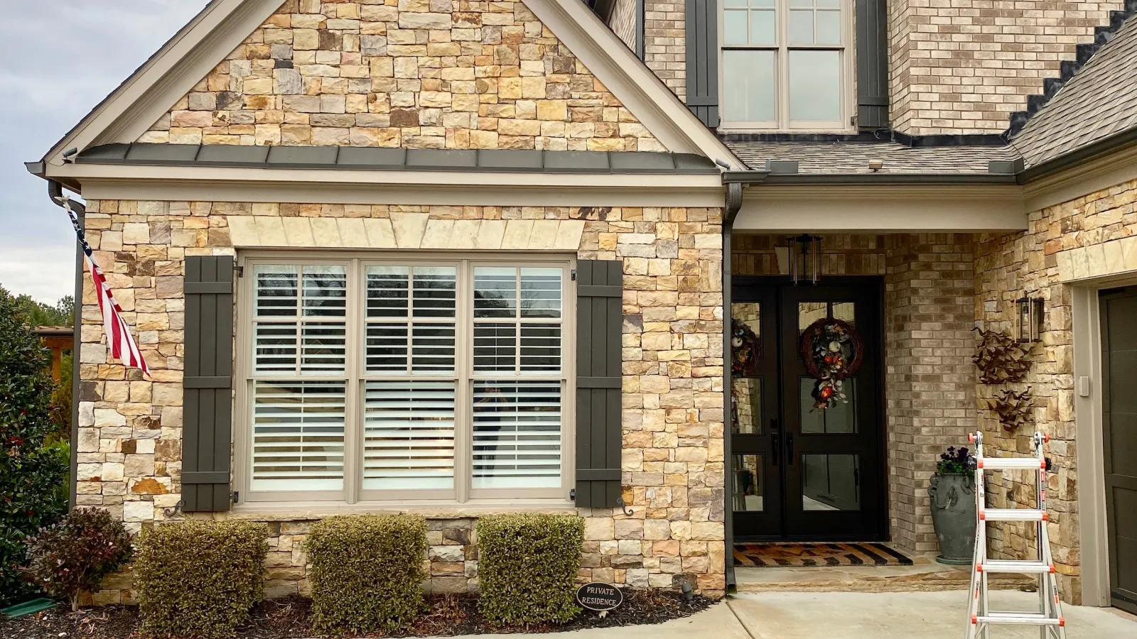 Front view of a modern two-story house with beige stone and brick exterior, black shutters, and a double black front door.