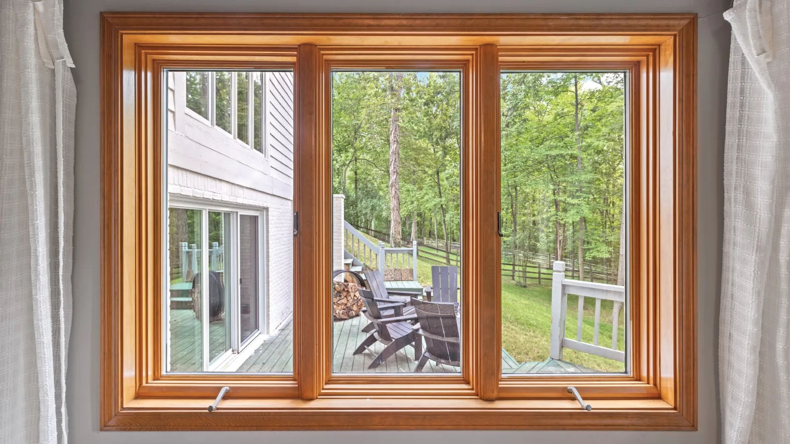 Large wooden-framed window overlooking a deck with chairs, firewood, and a green forested backyard.