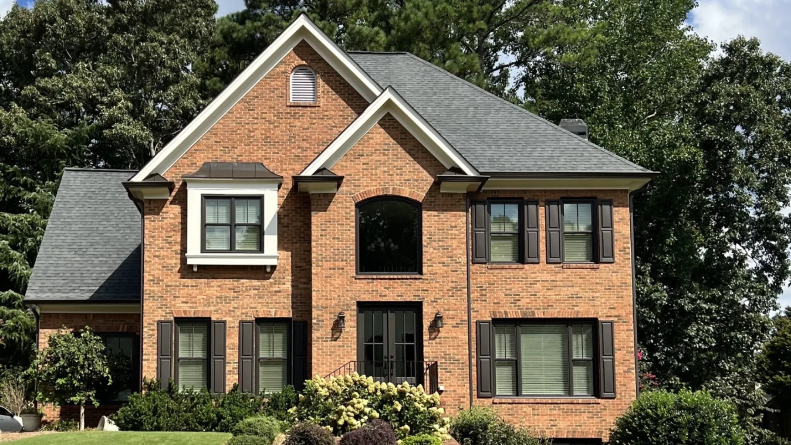 Two-story brick house with gray roof, black shutters, and landscaped garden under a blue sky with clouds.