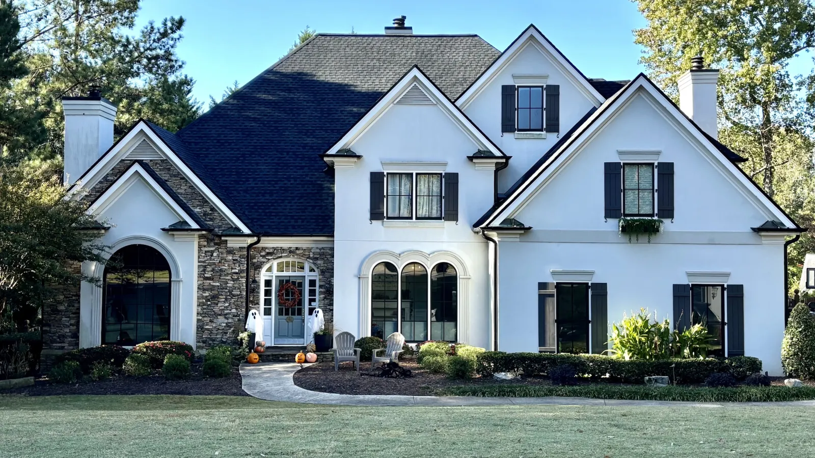 Large white two-story house with dark shutters, stone accents, and a manicured front lawn on a sunny day