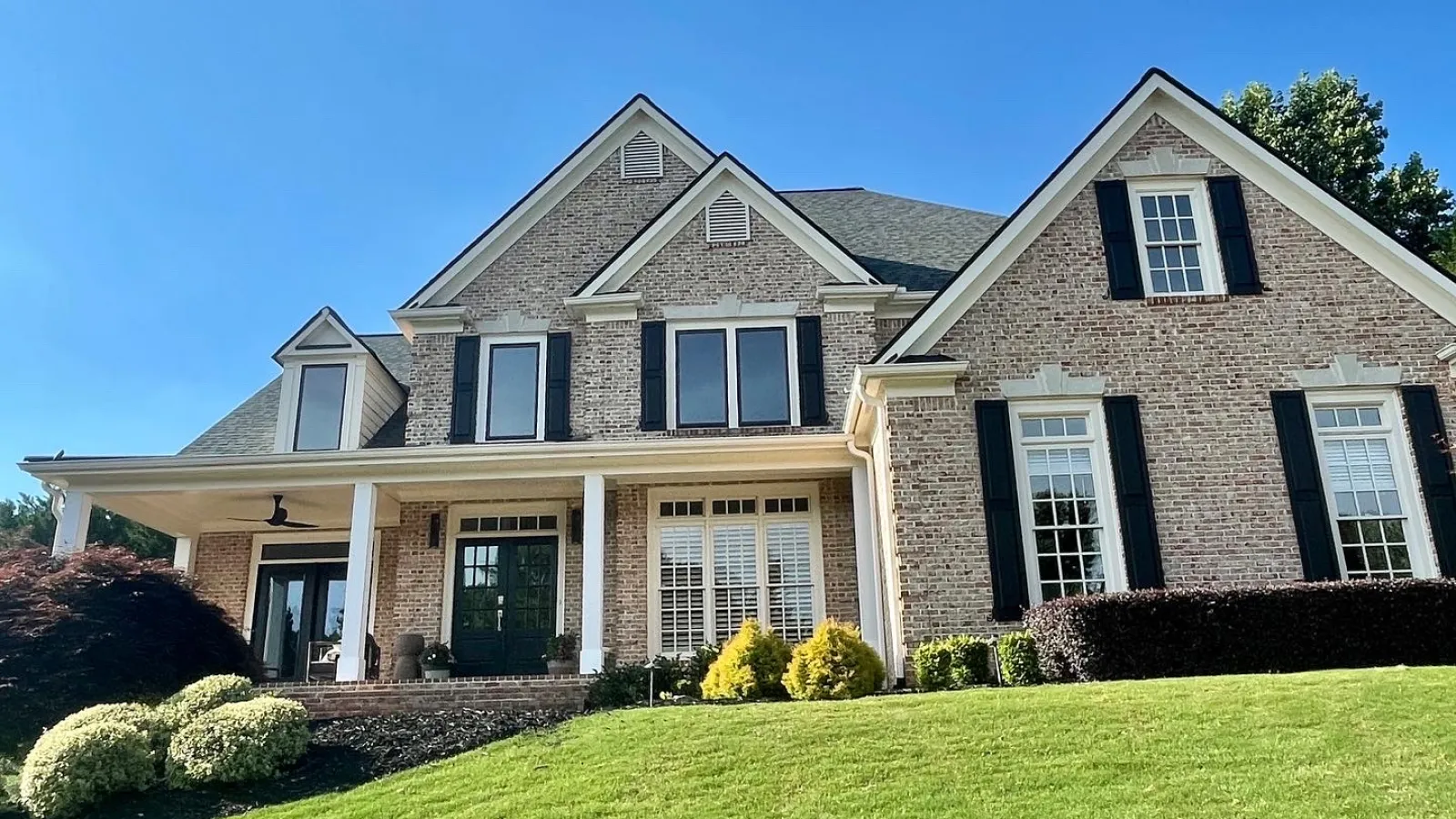Large brick house with black shutters, multiple gables, a covered porch, and a well-kept green lawn under clear blue sky
