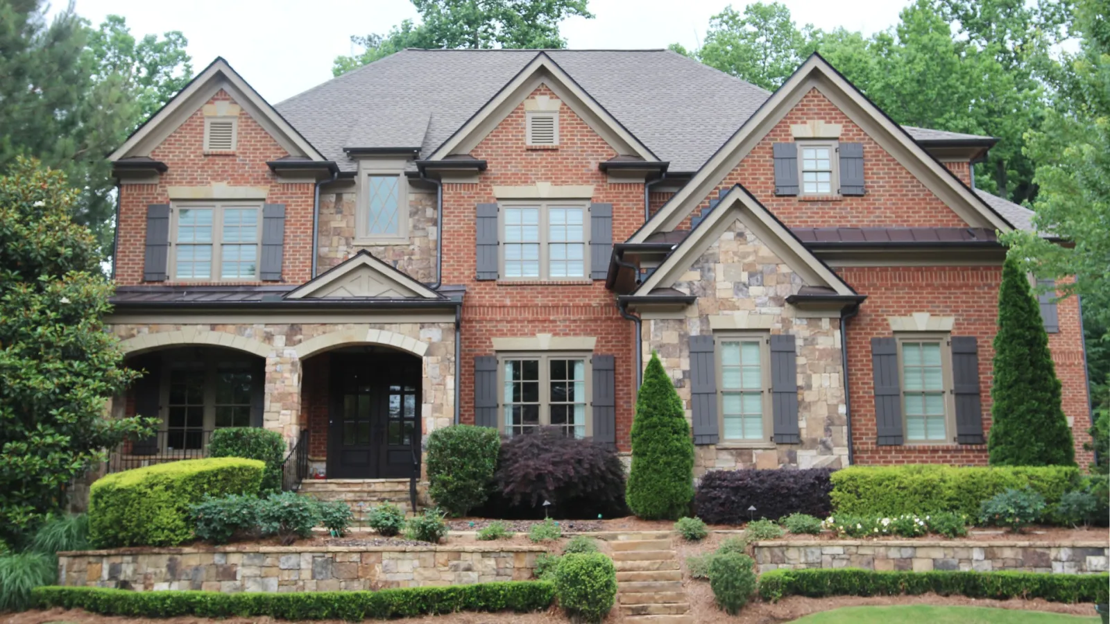 Two-story brick and stone house with multiple gables, arched entryway, and manicured shrubs and trees.