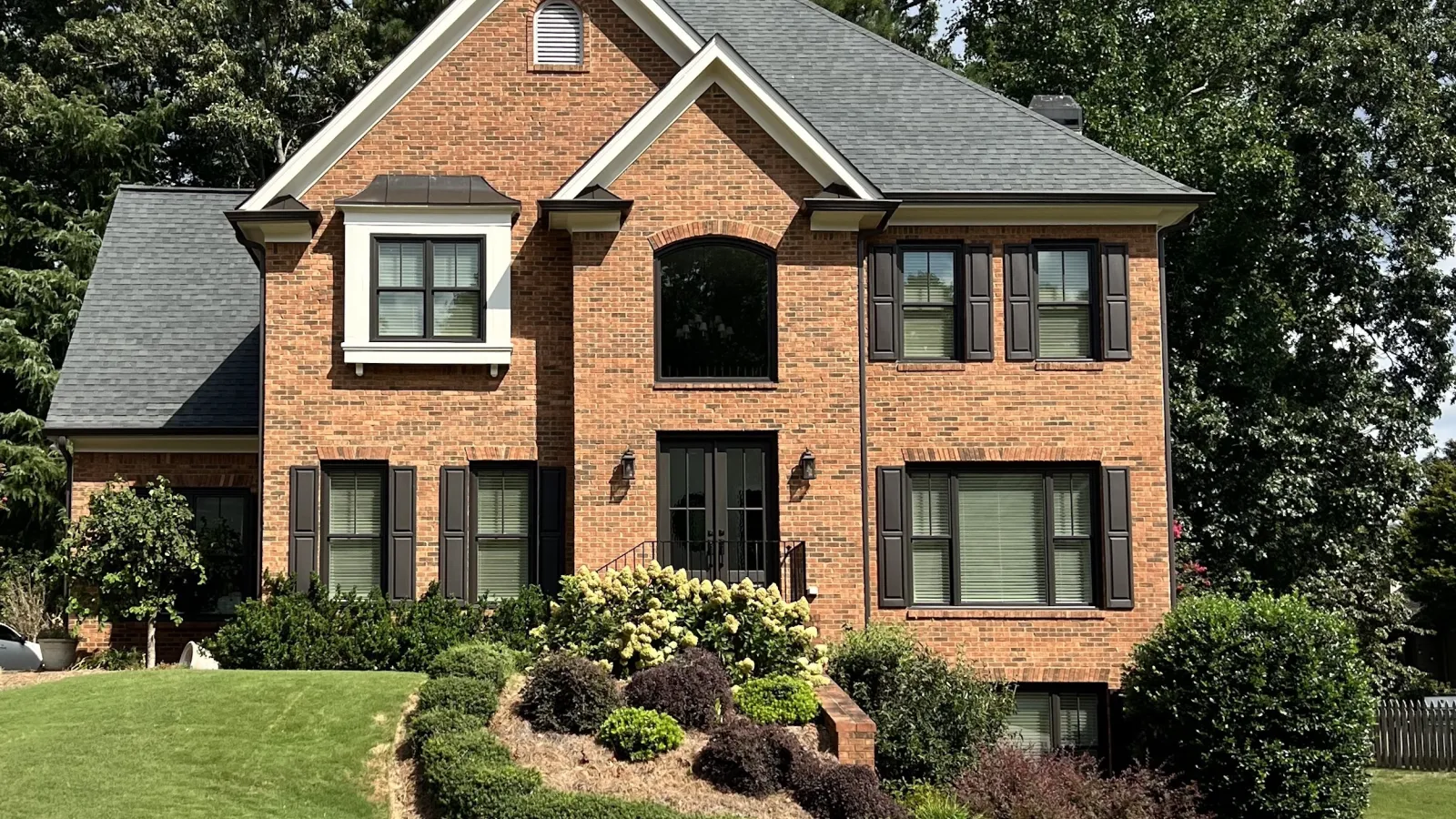 Two-story brick house with black shutters and landscaped front yard under blue sky with clouds.