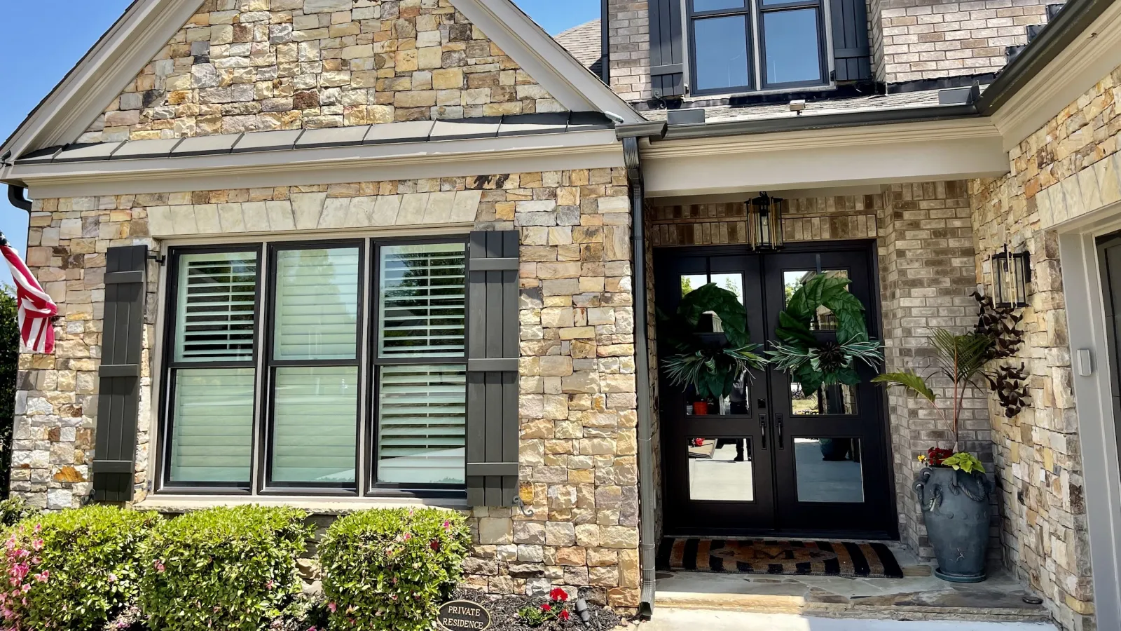 Stone and brick house exterior with black shutters, large windows, double front doors with green wreaths, and landscaping.