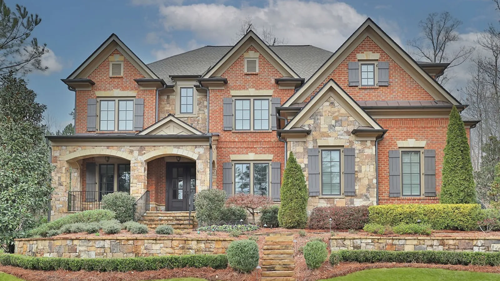 Large two-story brick and stone house with multiple windows, shutters, and landscaped front yard under blue sky.