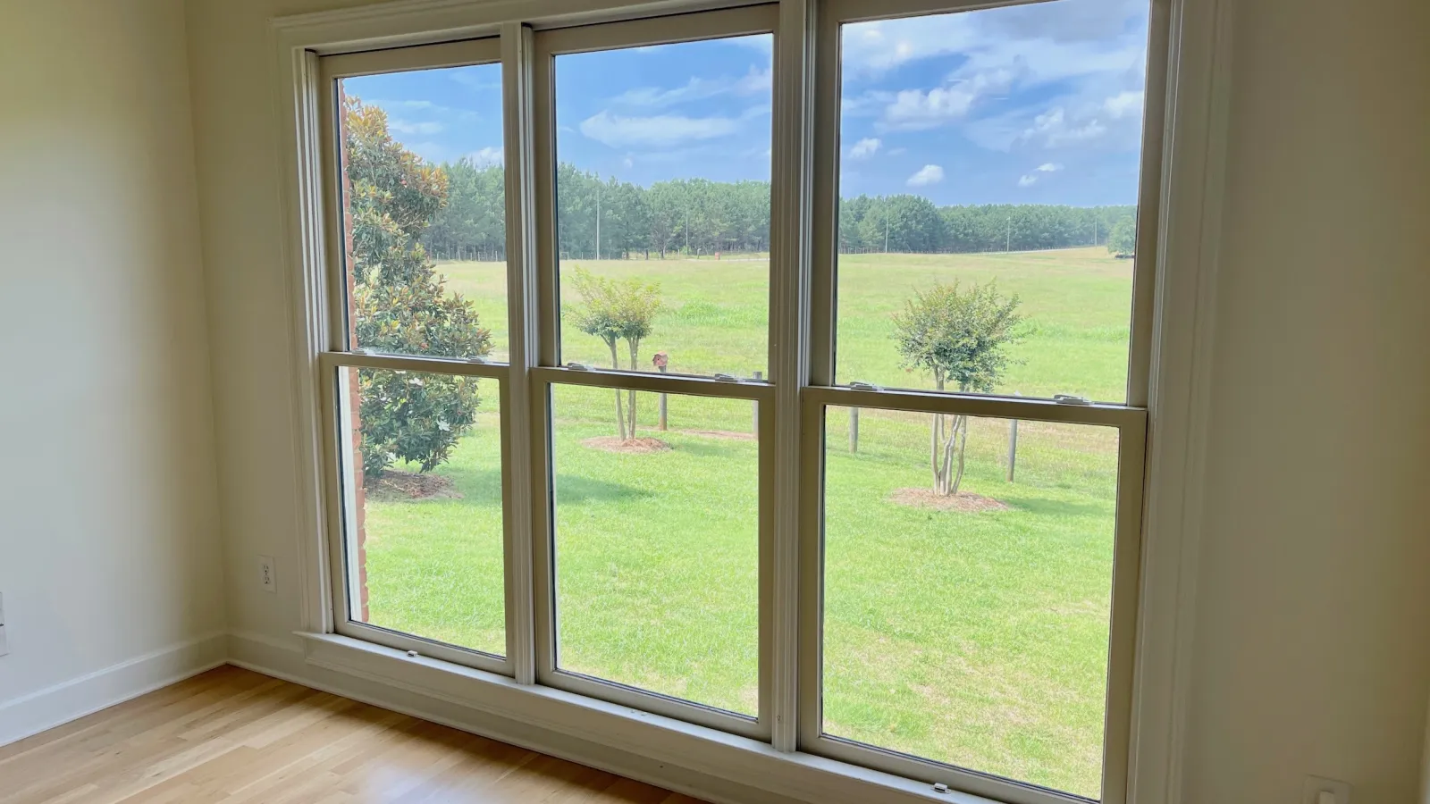 Large triple-pane window overlooking green lawn with small trees and blue sky in empty room with hardwood floor.
