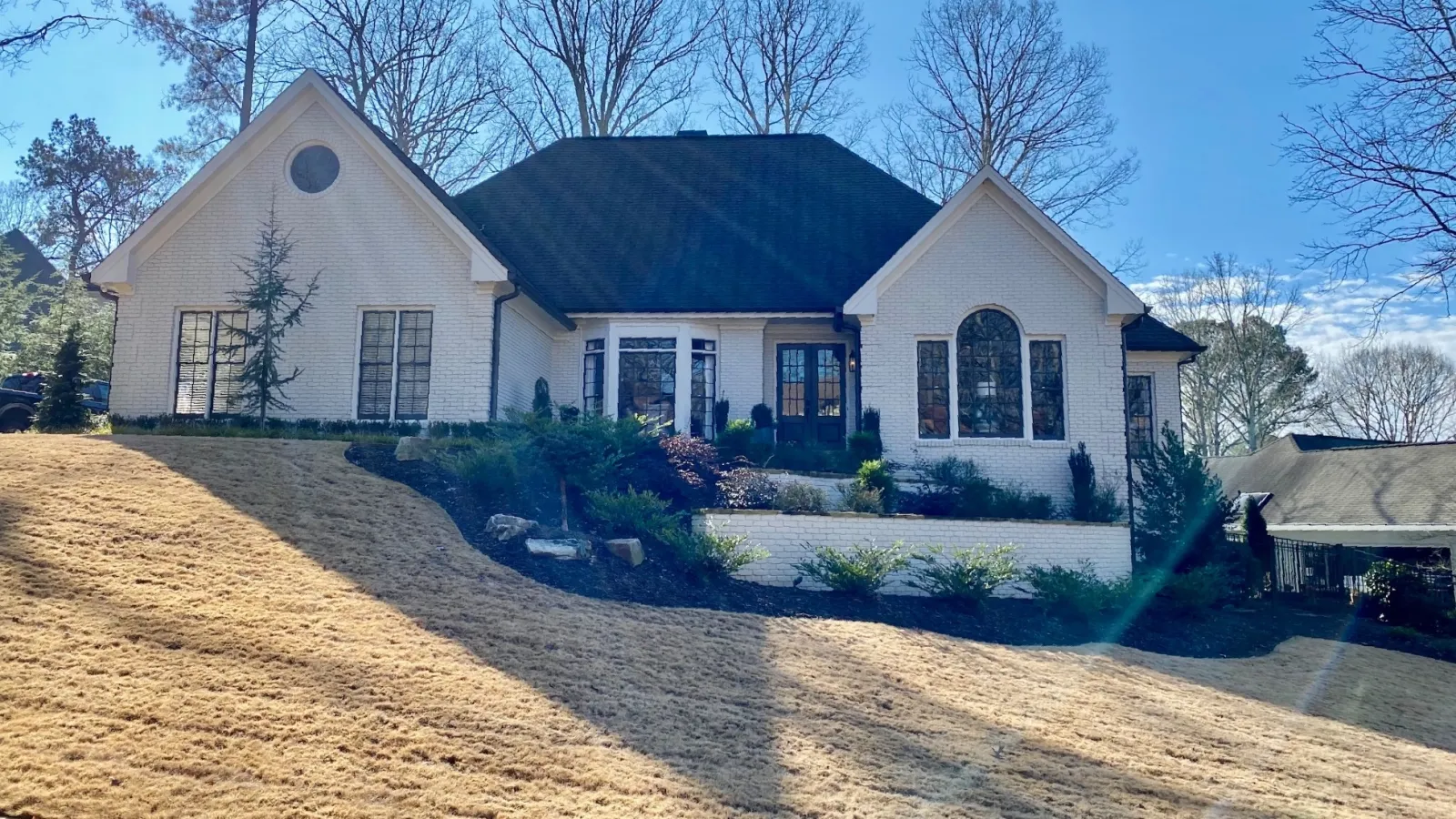 Two-story white brick house with black roof on a sloped lawn under a clear blue sky with leafless trees.