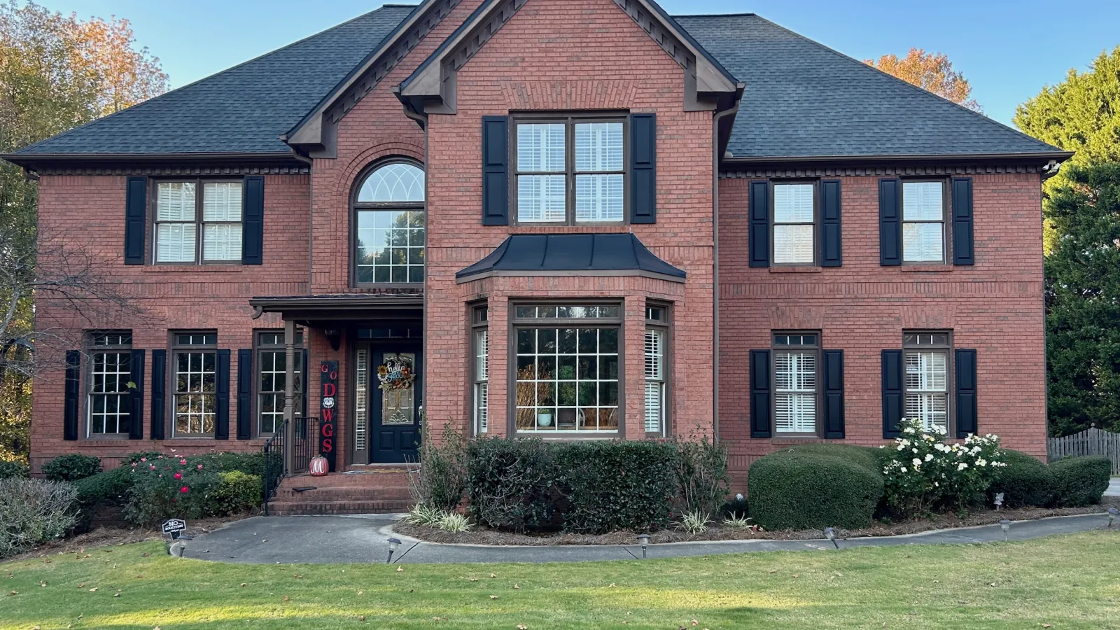 Two-story red brick house with black shutters, large front lawn, and decorative entrance door with wreath.