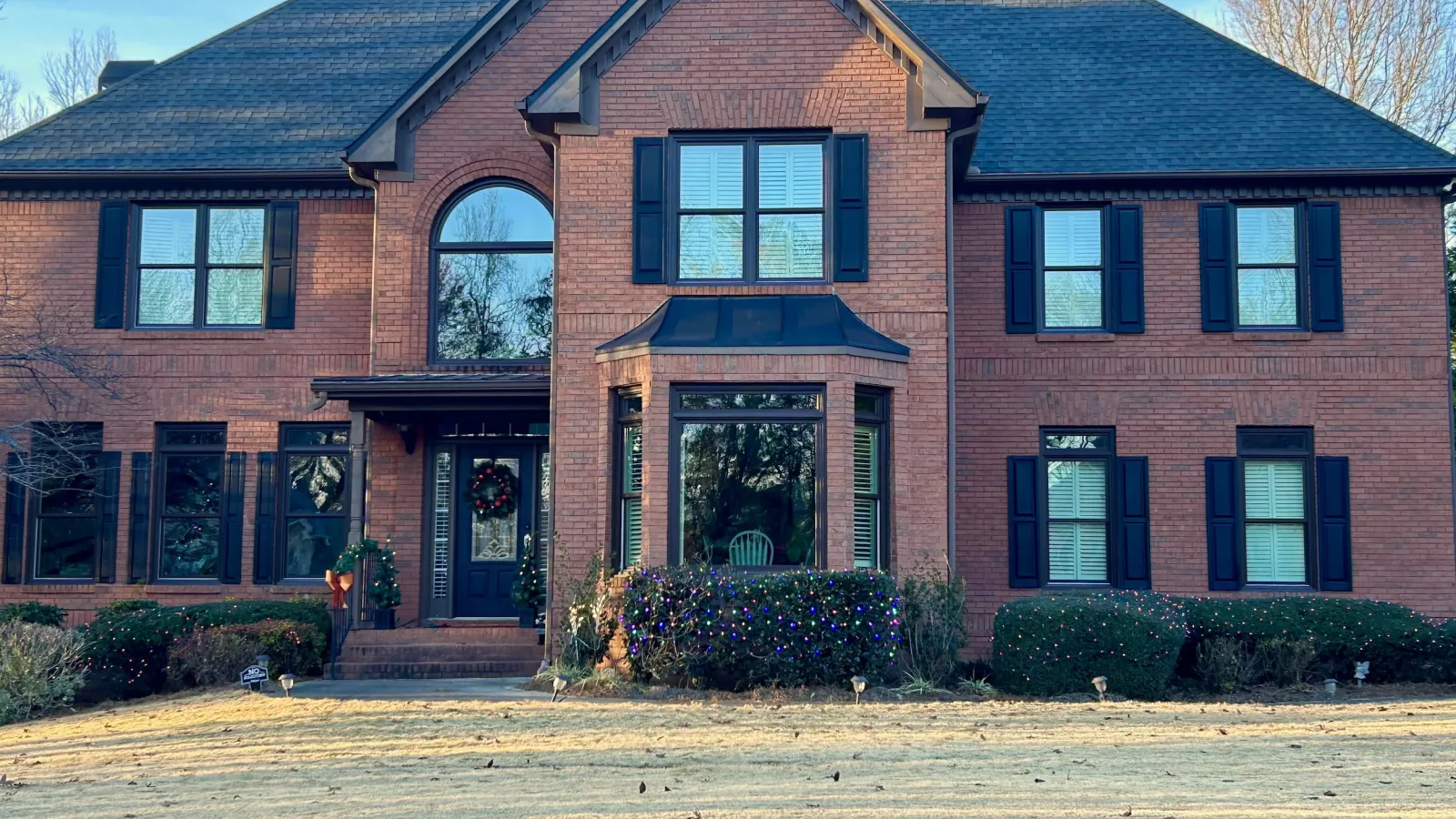 Two-story brick house with black shutters, large windows, and a decorated front door in winter daylight.
