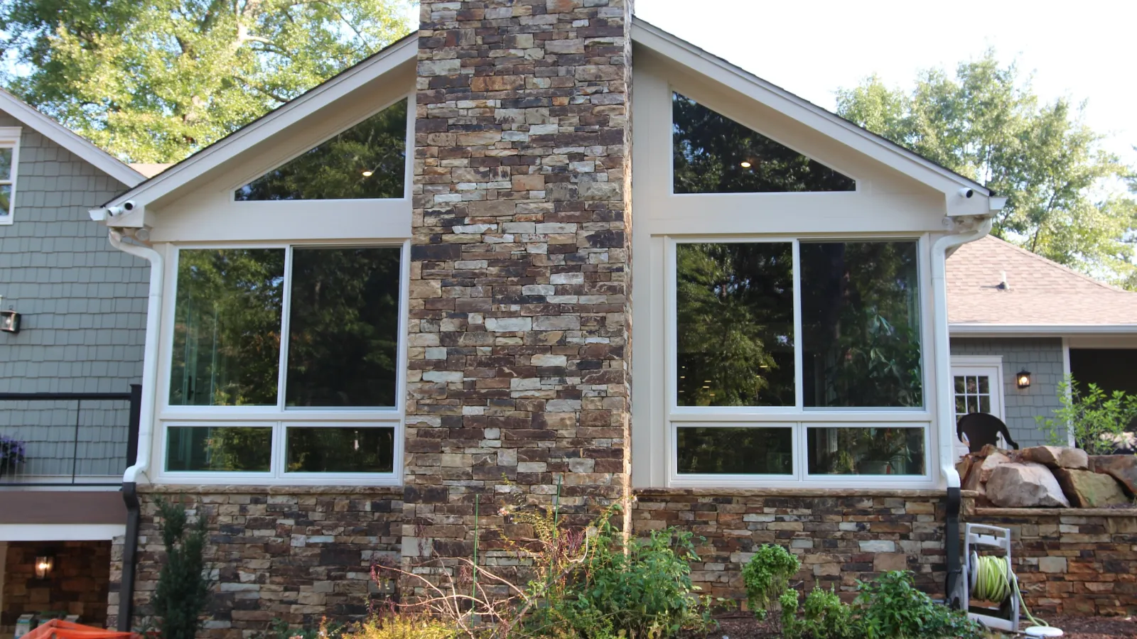 House exterior with large symmetrical windows and a central stone chimney surrounded by green plants and trees.