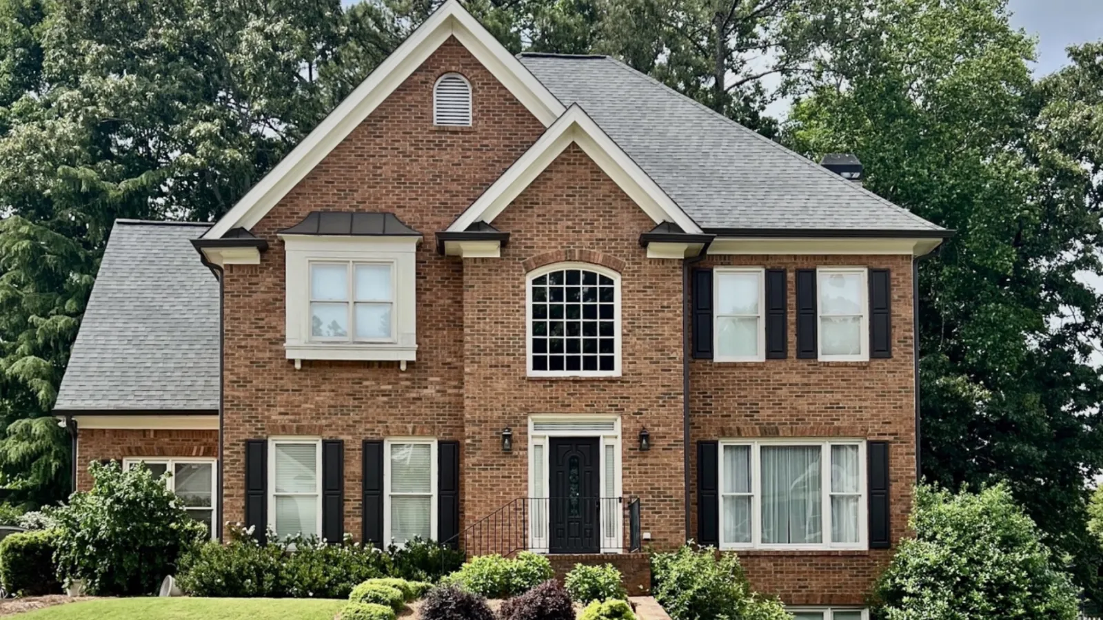 Two-story brick house with black shutters, manicured lawn, and landscaped garden under cloudy sky.