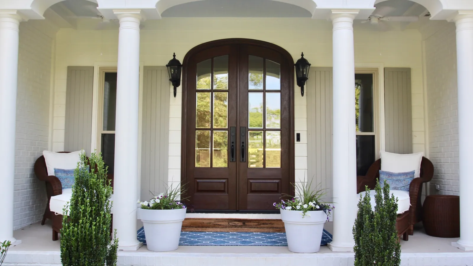 Front porch with double wooden doors, white columns, wicker chairs, potted plants, and blue rug under a covered entry