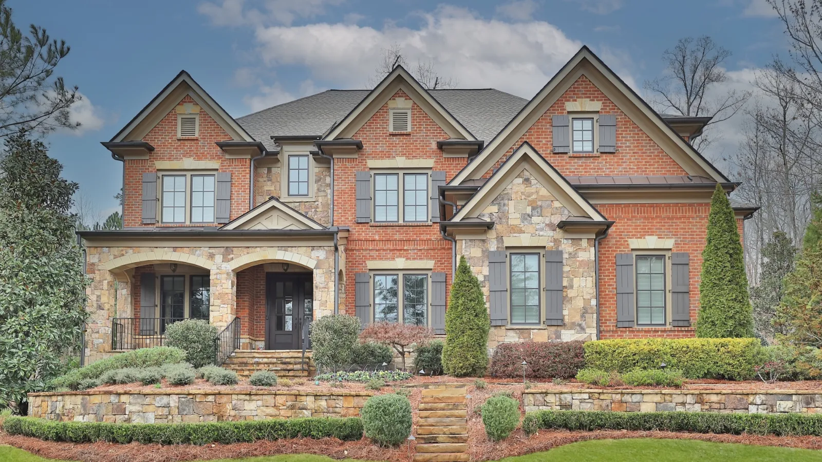 Large two-story brick and stone house with manicured lawn, front steps, shrubs, and trees under a partly cloudy sky
