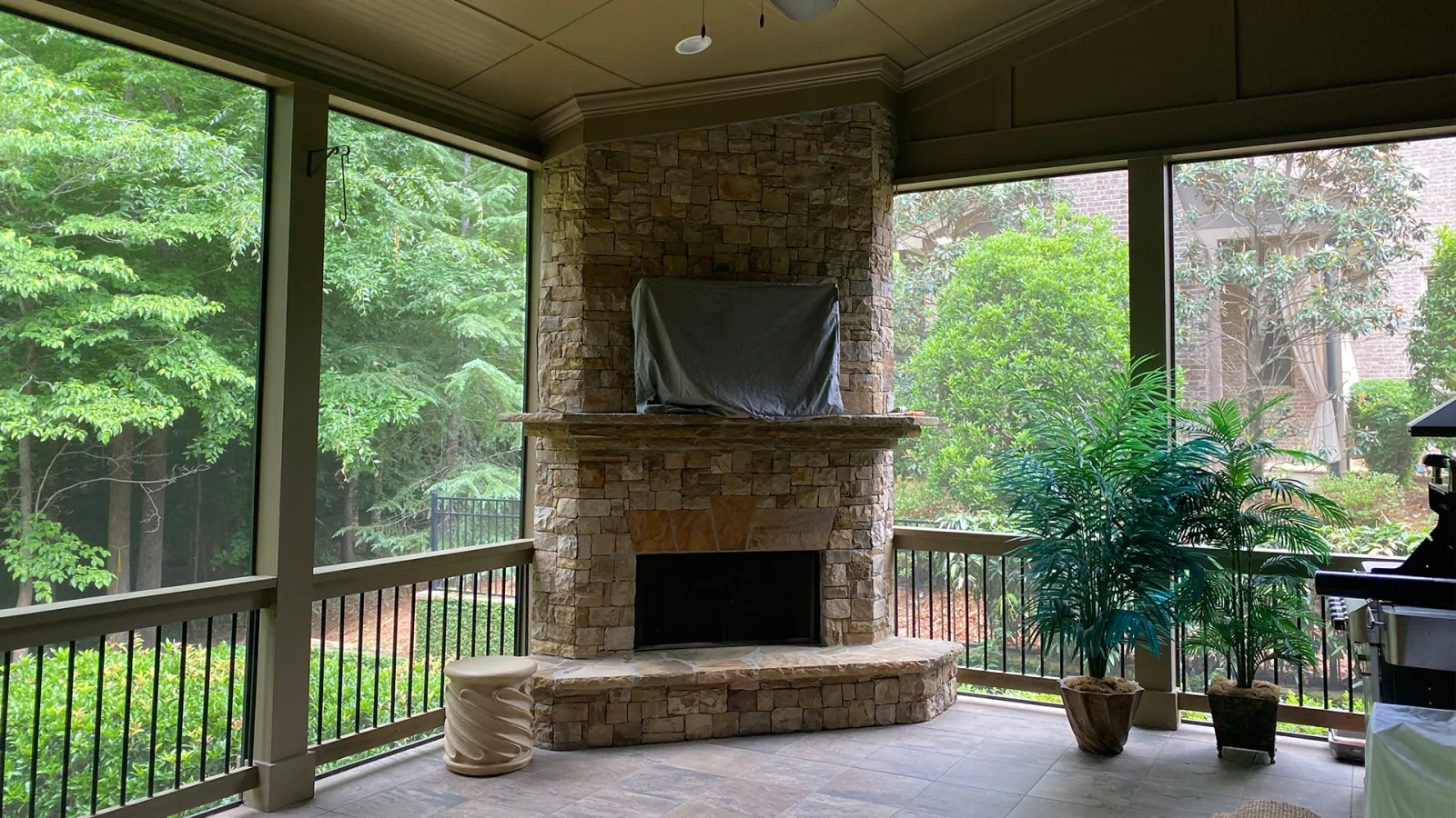 Covered outdoor patio with stone fireplace, potted plants, and views of green trees in a tranquil backyard.