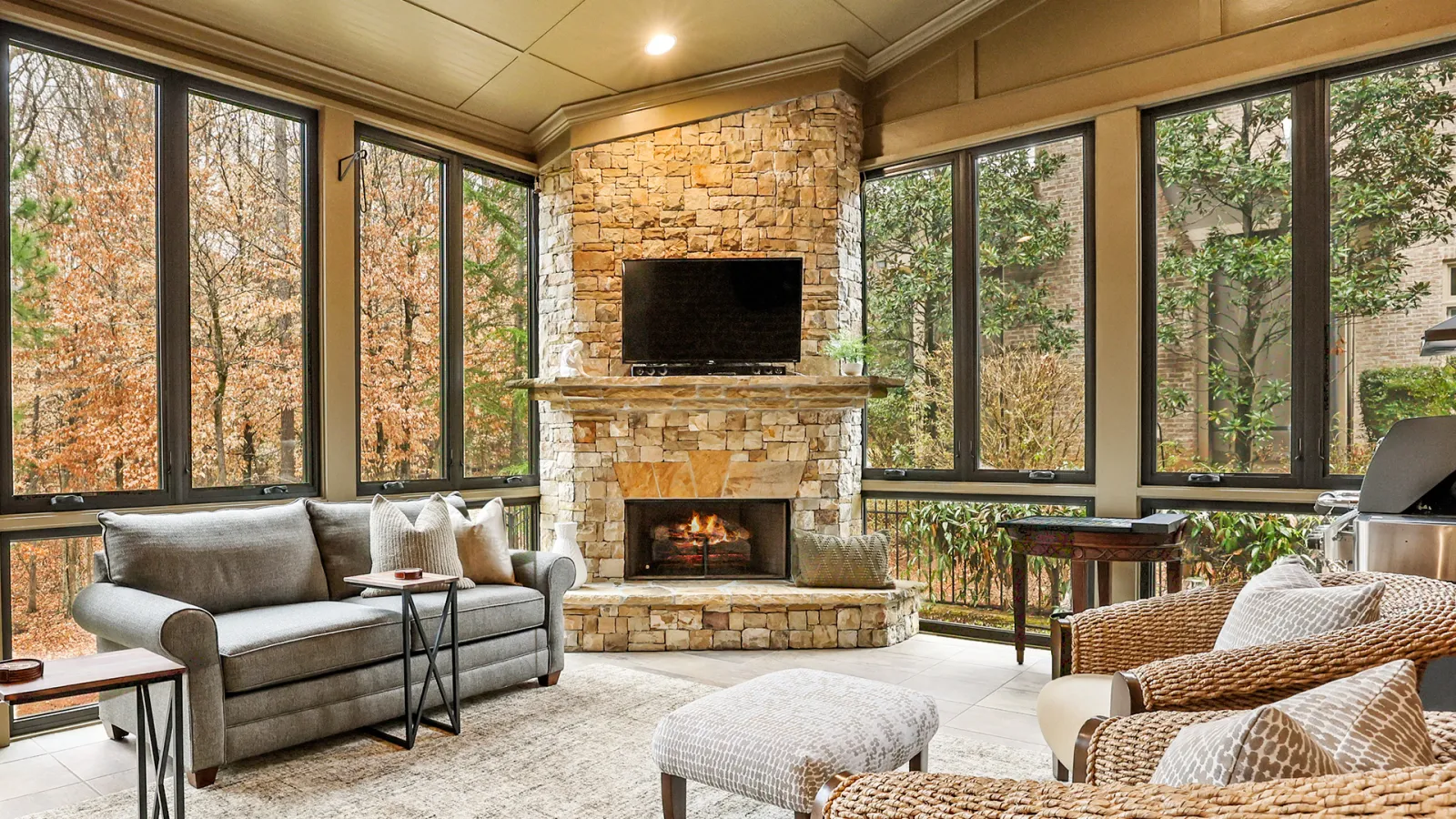 Cozy sunroom with stone fireplace, gray sofa, wicker chairs, large windows showing autumn foliage outdoors.