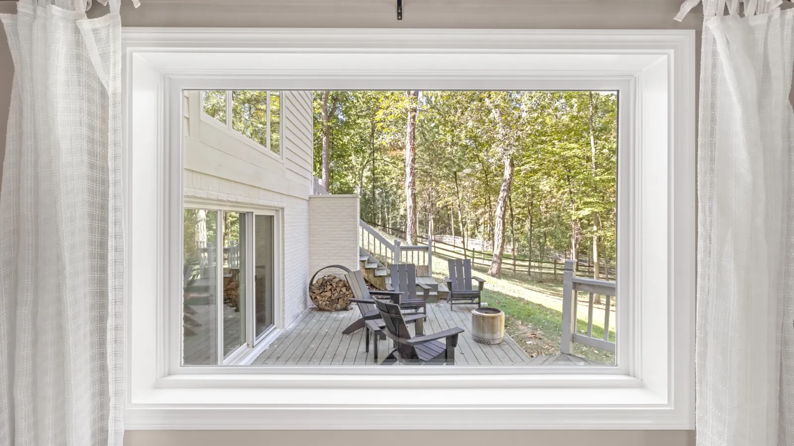 View through a large window of a wooden deck with Adirondack chairs and forest in the background.