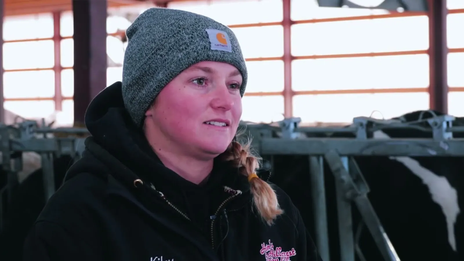 Young woman wearing a gray beanie and black jacket inside a barn with cows in the background.