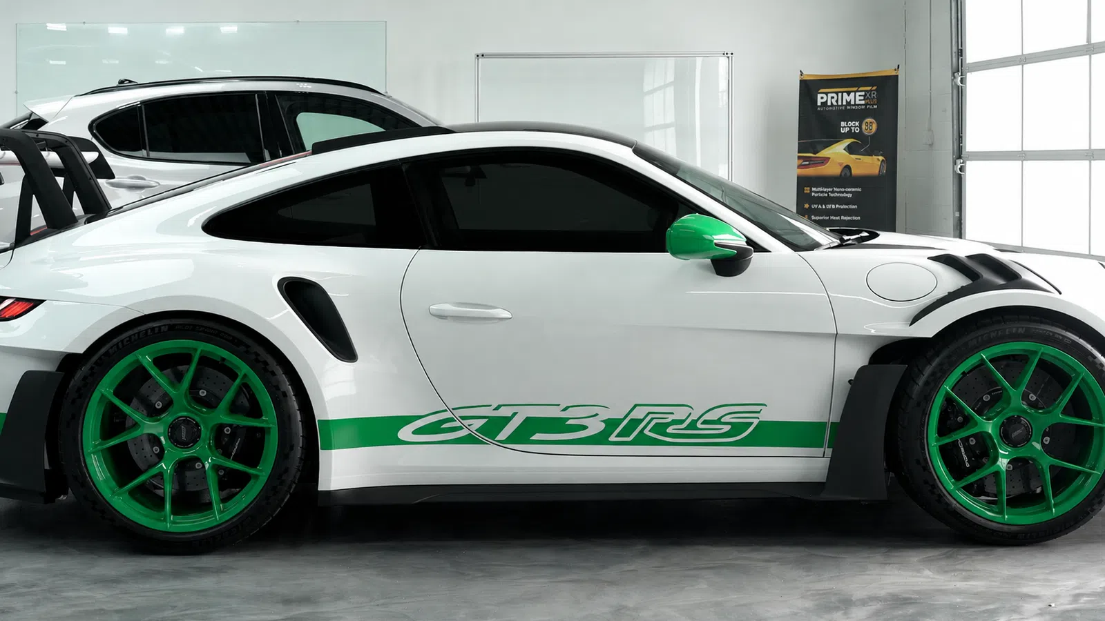 White Porsche GT3 RS sports car with green wheels and side decals in modern garage setting.