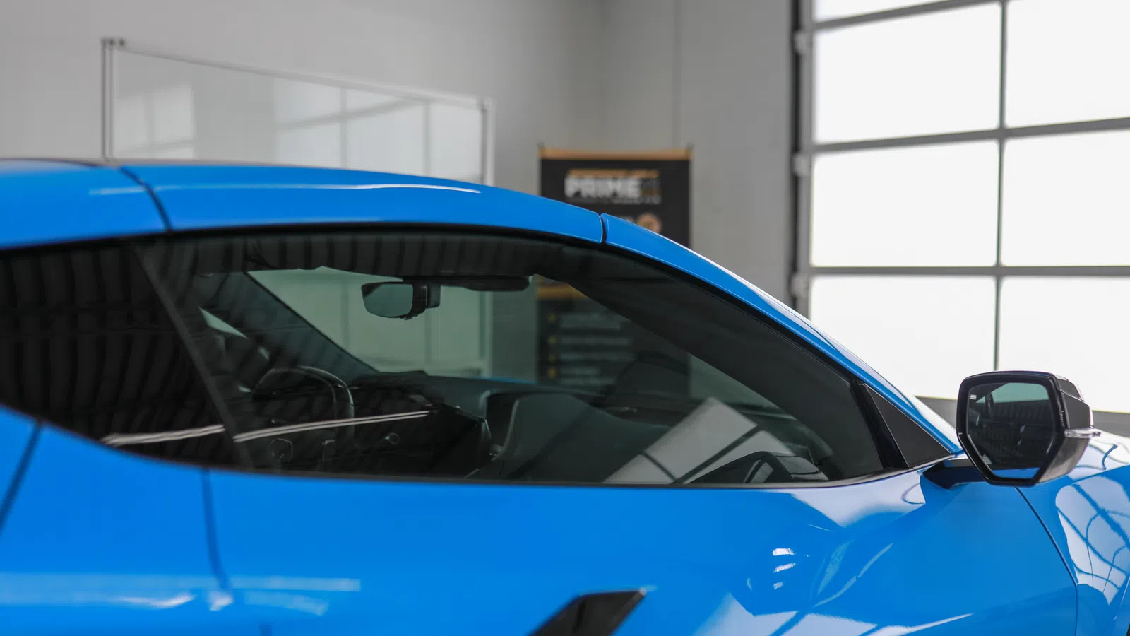 Close-up of a blue sports car door and window inside a bright garage with large windows.