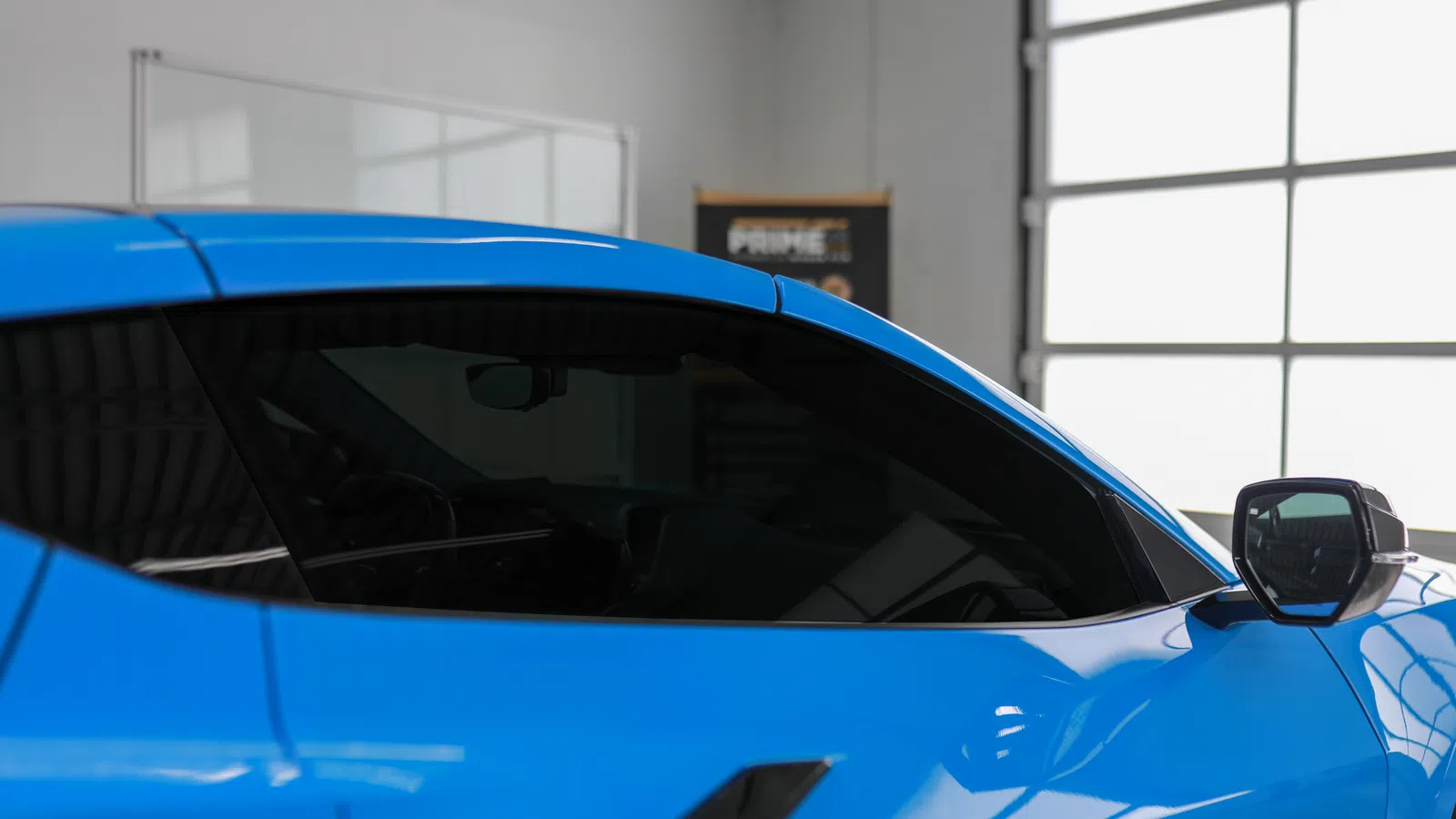 Close-up of a vibrant blue sports car side showing tinted windows and sleek door mirror in a bright garage.