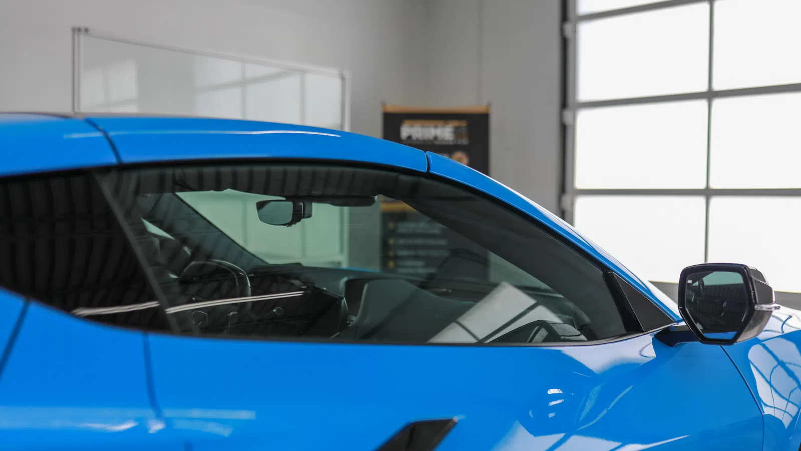 Close-up of a bright blue sports car's side window and mirror inside a modern garage with large windows.