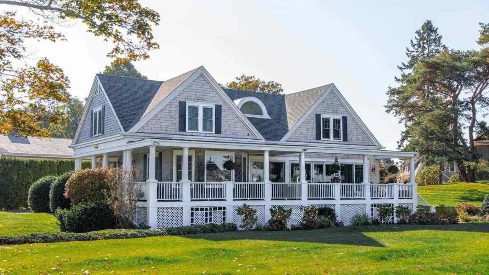 Large white porch house with gray shingles, black shutters, and manicured lawn surrounded by trees in fall.