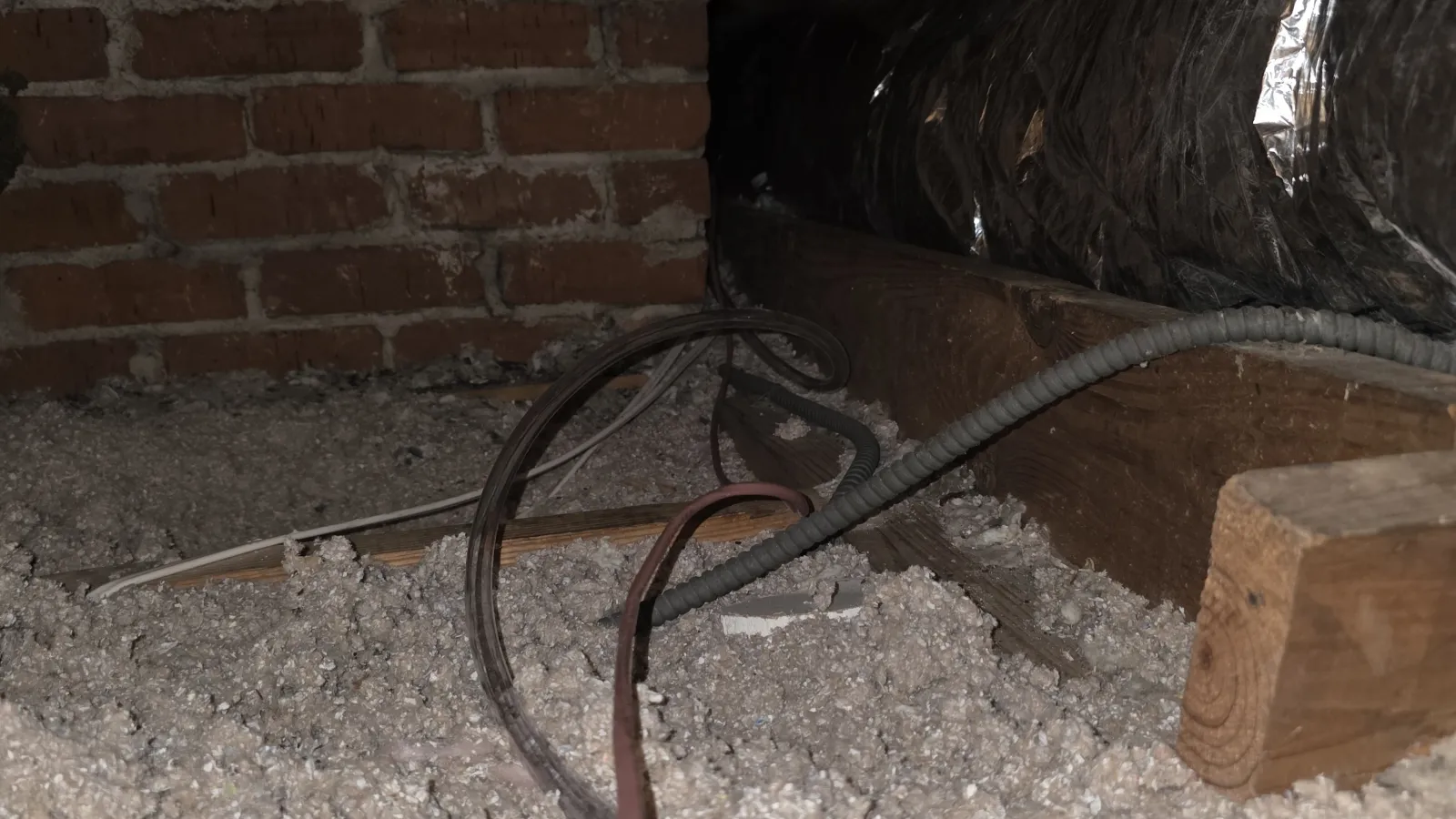 Insulated attic space with ducts, wooden beams, brick wall, and electrical wiring over blown-in insulation.