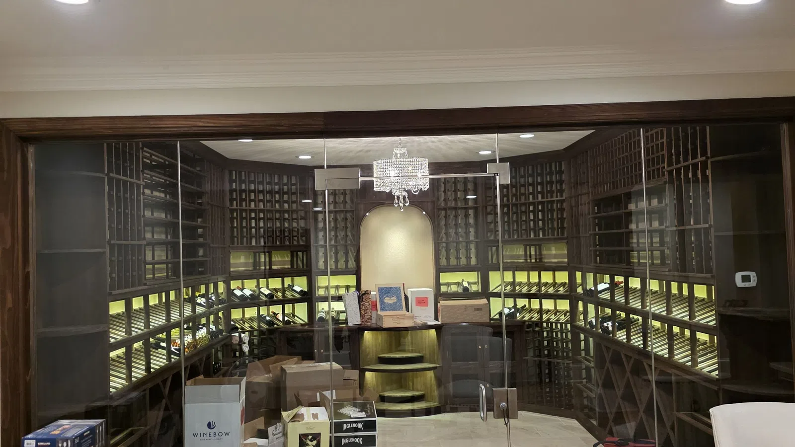Spacious wine cellar with wooden racks, glass doors, cardboard boxes, and a chandelier overhead.
