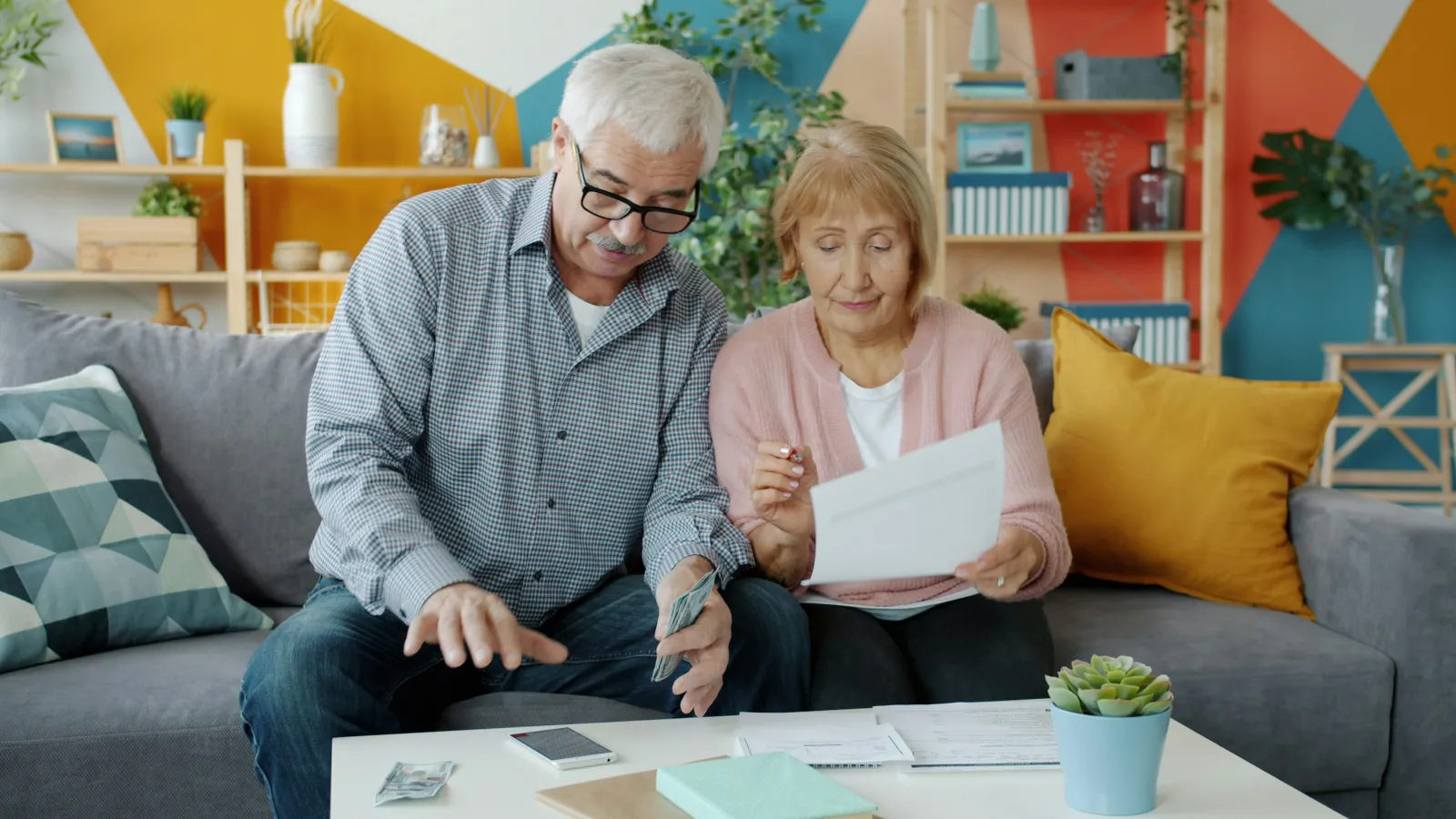Senior couple sitting on sofa reviewing bills and counting cash in a colorful living room with plants.