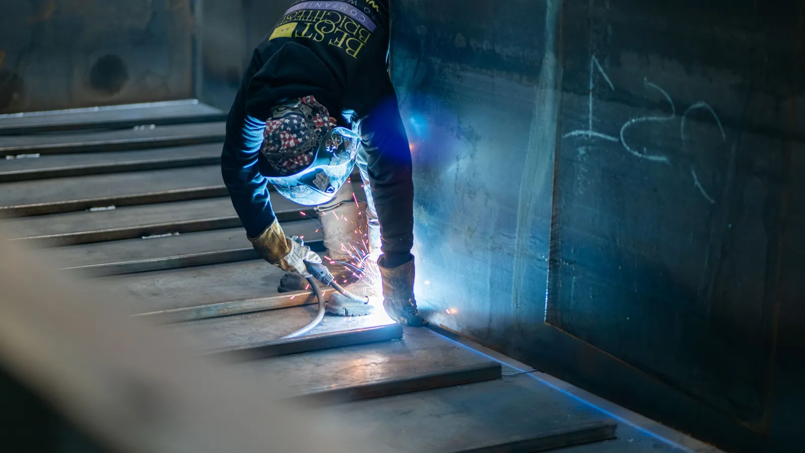 Welder wearing protective gear sparks metal welding inside industrial workshop environment.