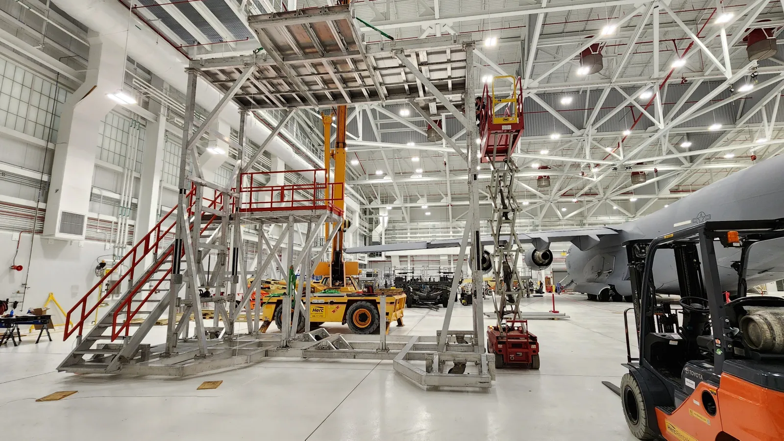 Aircraft maintenance hangar with scaffolding, platforms, forklifts, and a military jet under bright industrial lighting.