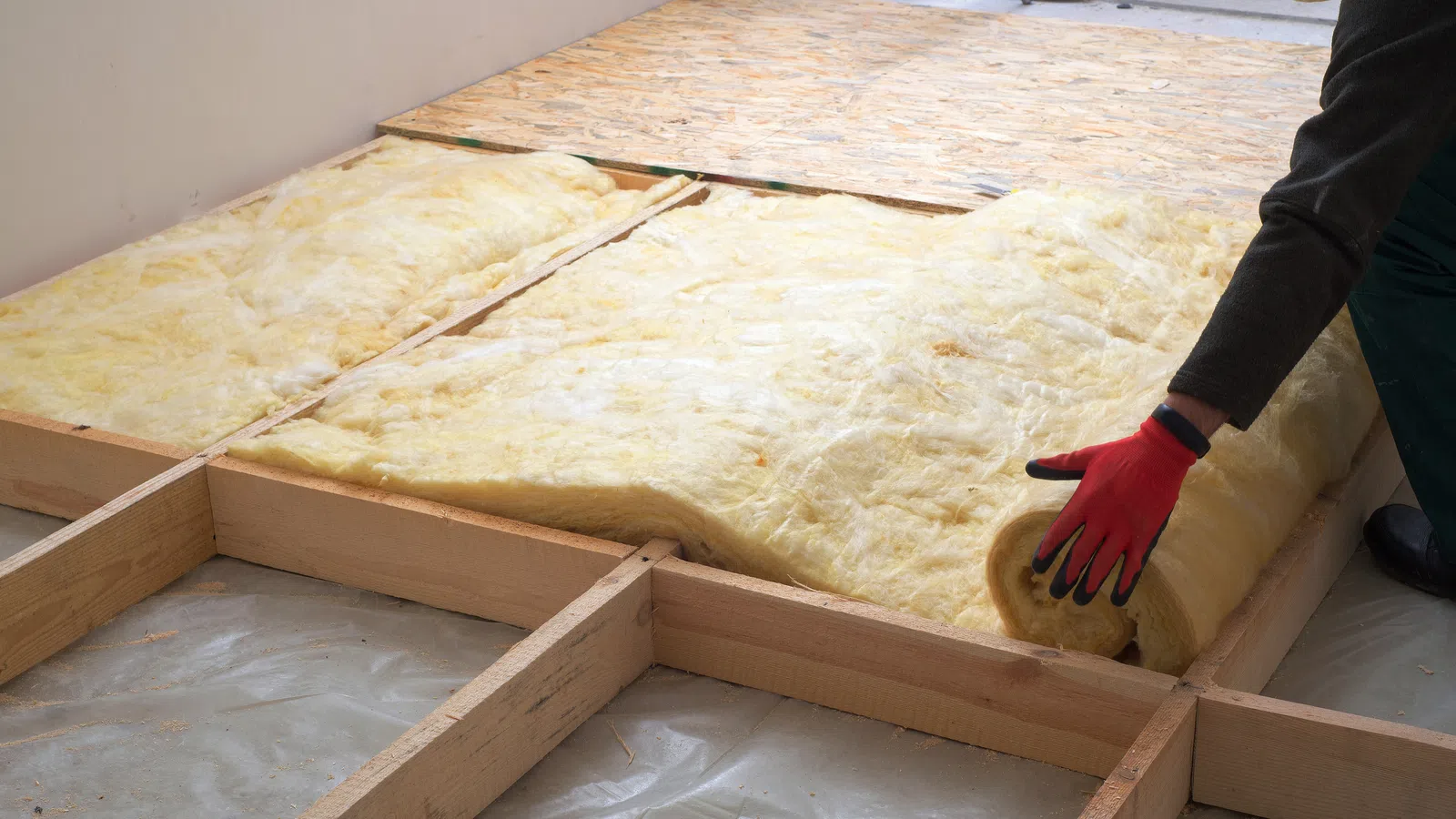 Worker installing yellow fiberglass insulation rolls in wooden floor framework during home construction.