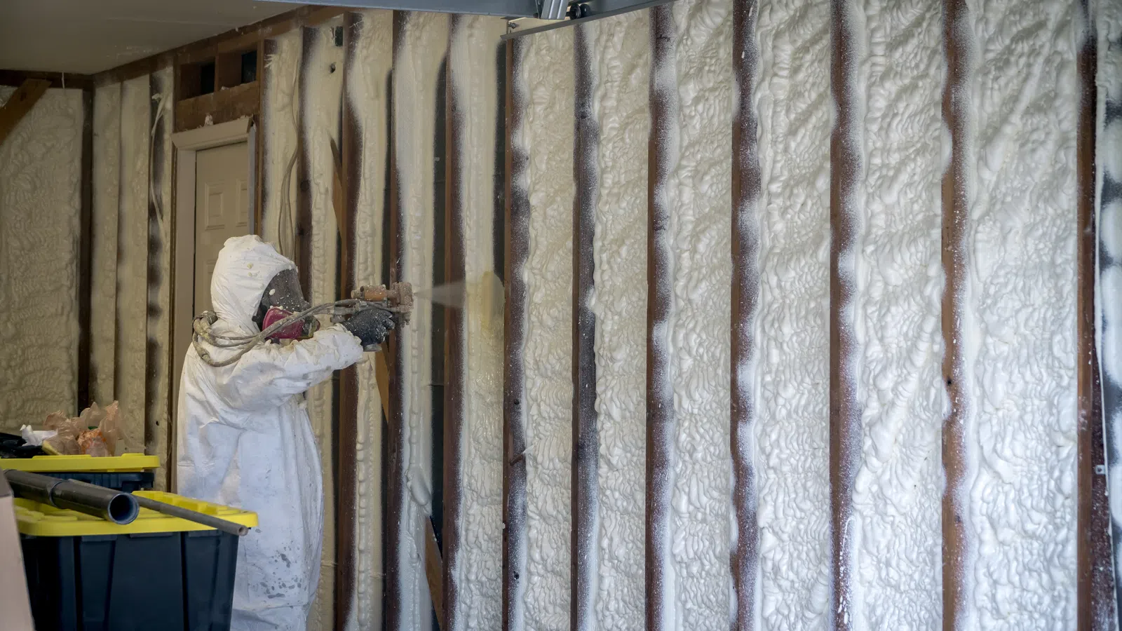Worker in protective gear spraying foam insulation on wall studs in an unfinished room interior.