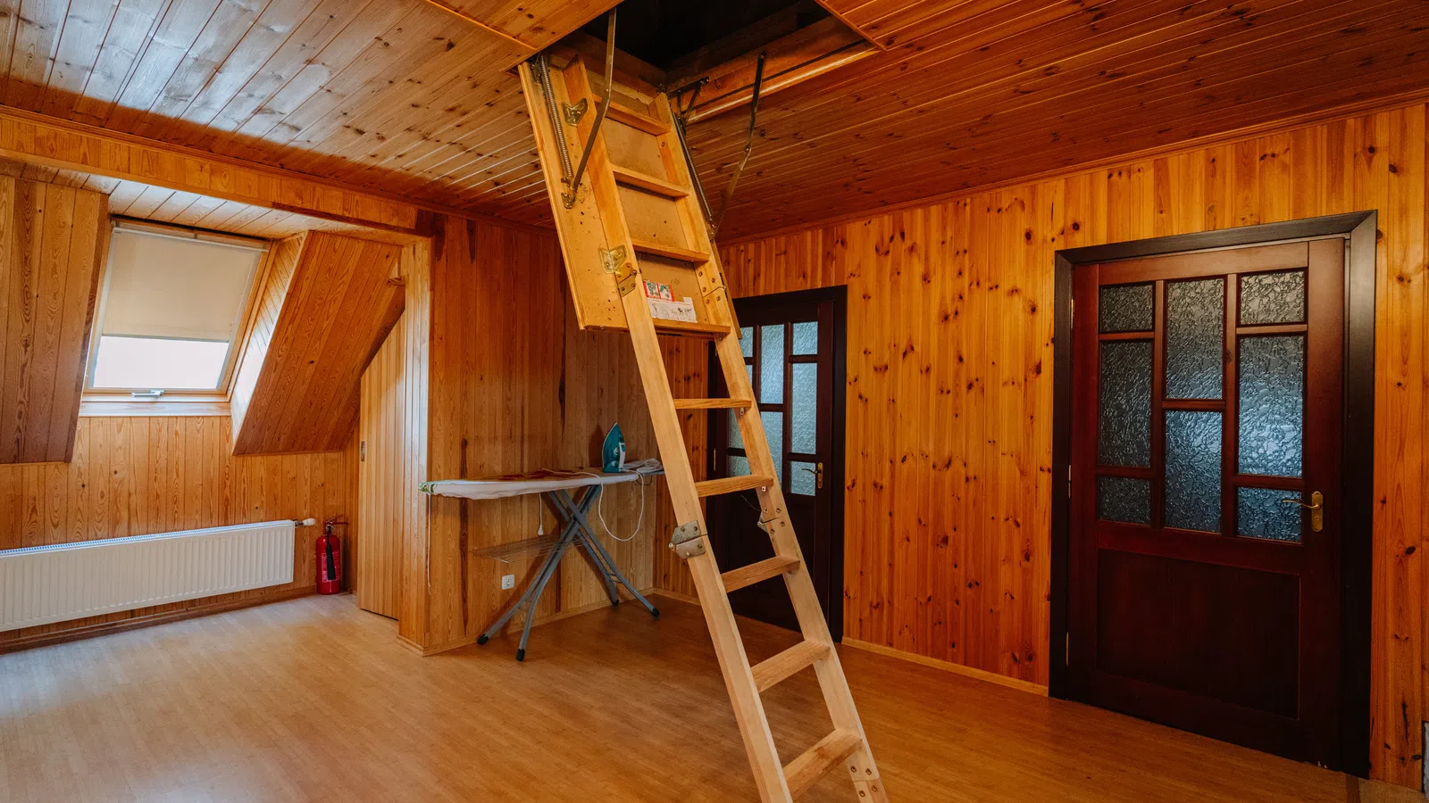 Wood-paneled attic room with foldable wooden ladder leading to ceiling hatch and ironing board by window.