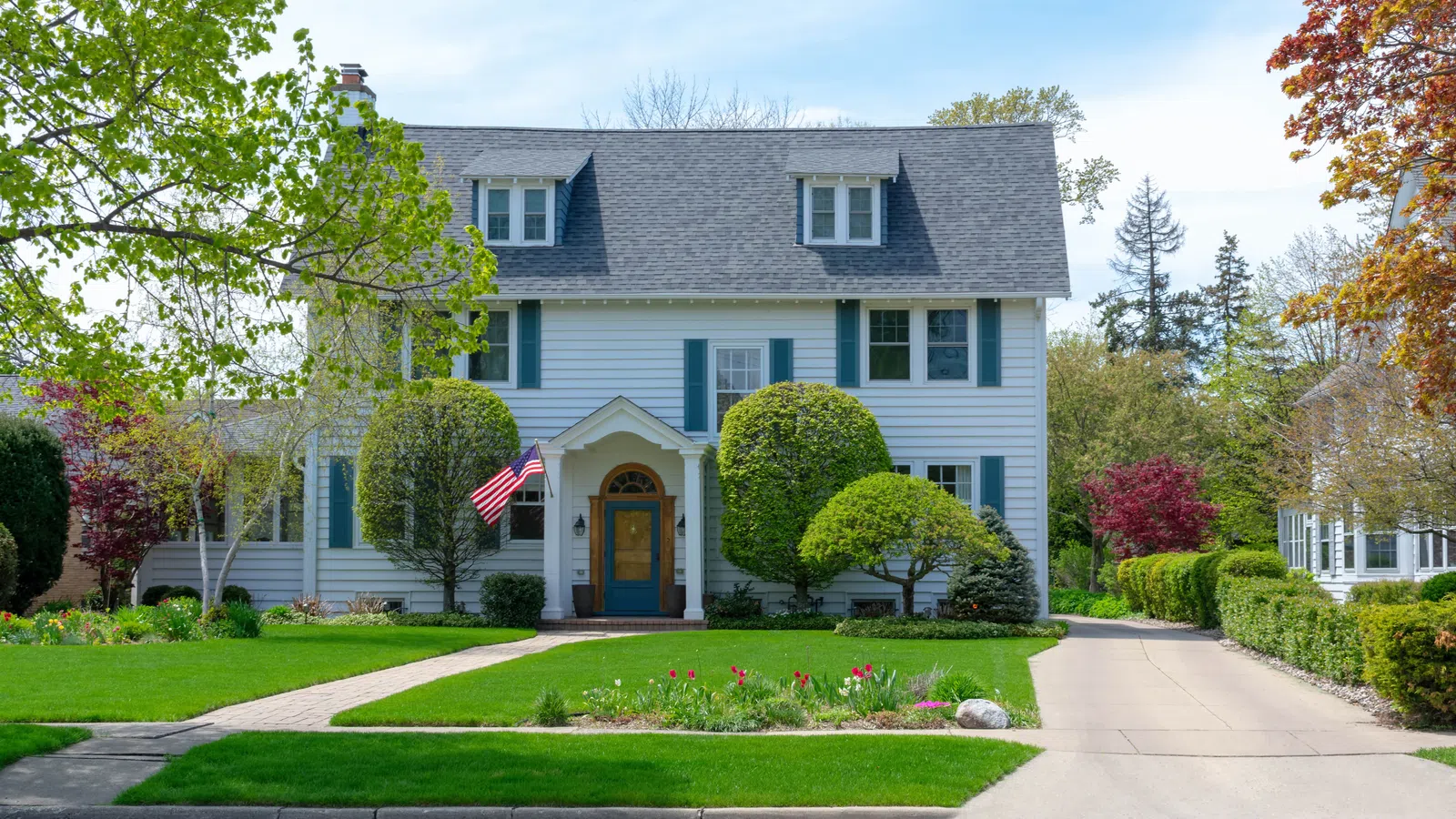 White two-story house with blue shutters, manicured lawn, trimmed bushes, paved driveway, and an American flag.