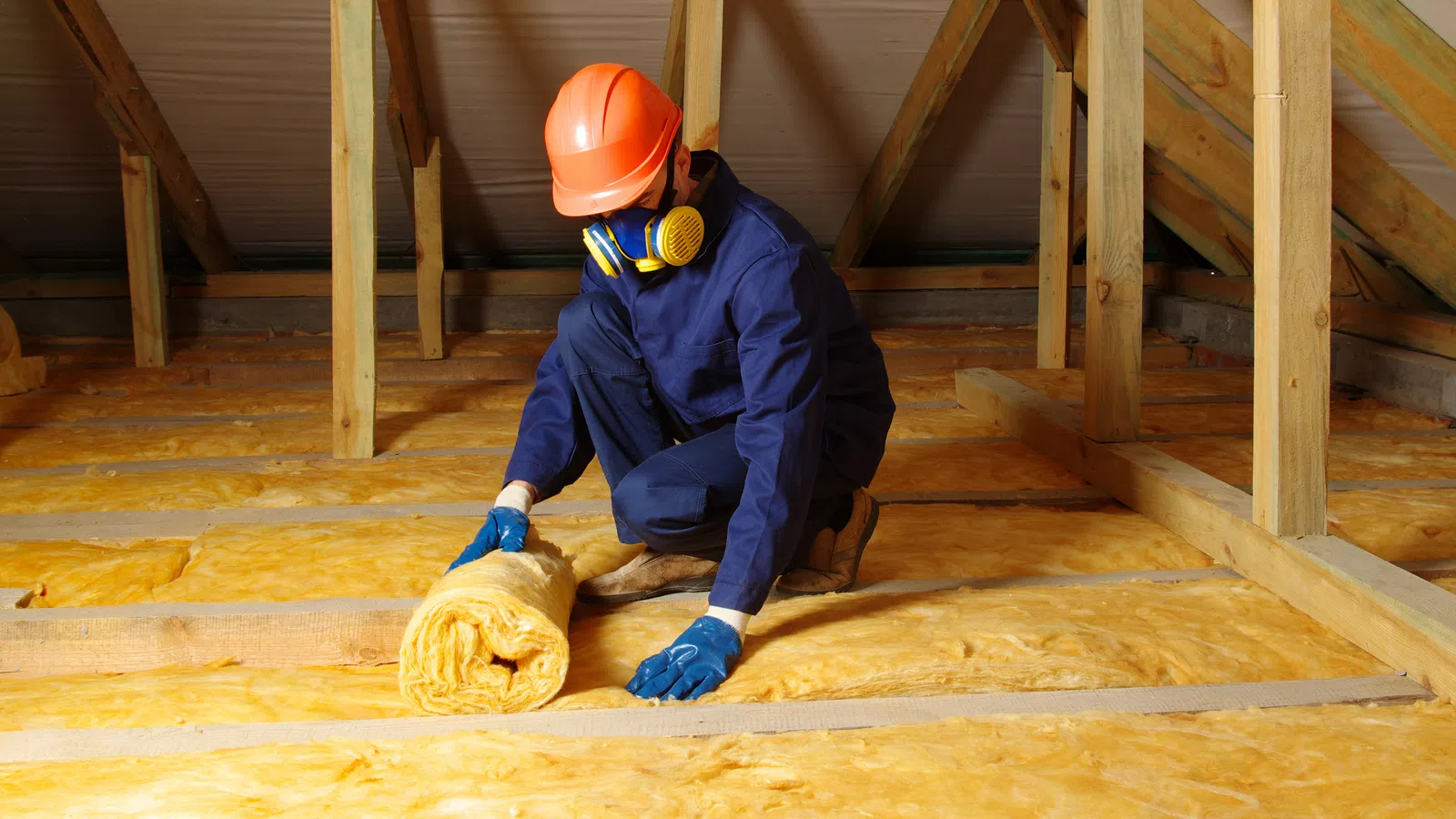 Worker in safety gear installing yellow fiberglass insulation in an attic space of a wooden framed house.