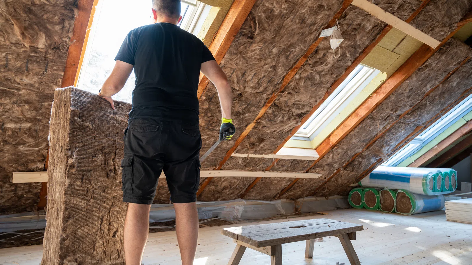 Man installing insulation in attic under a wooden roof with skylights and construction materials around.