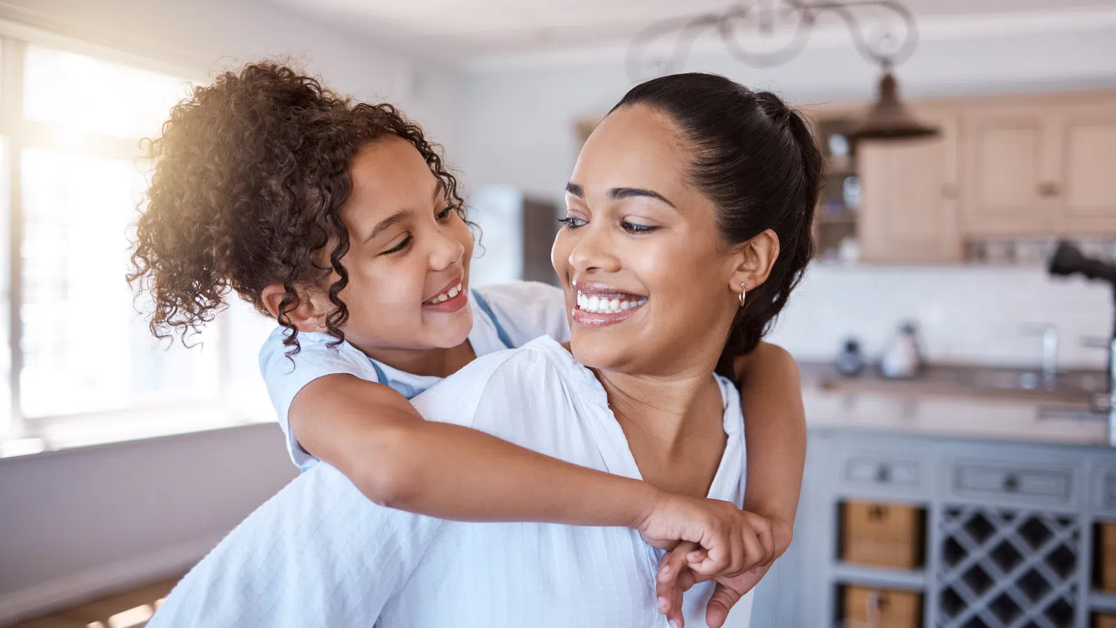 Smiling mother and daughter embracing joyfully in a bright, modern kitchen with natural sunlight.