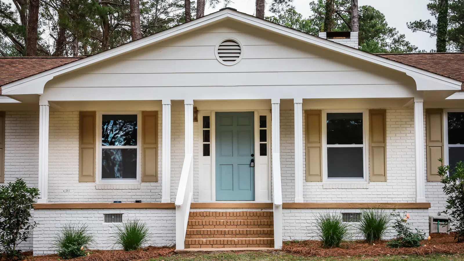 Front view of a white brick house with a blue door, beige shutters, porch, steps, and surrounding greenery.