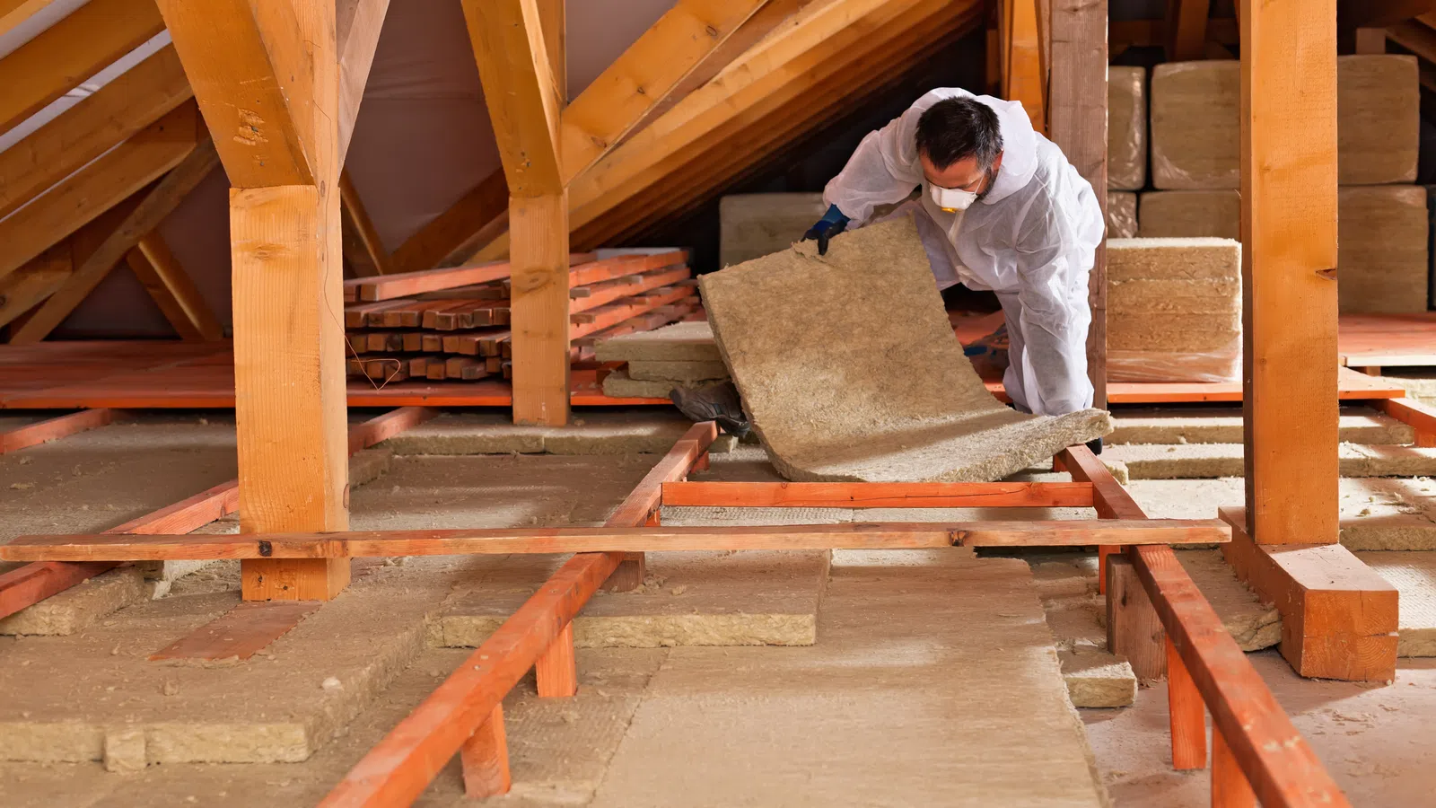 Worker in protective gear installing insulation panels in wooden attic space under roof