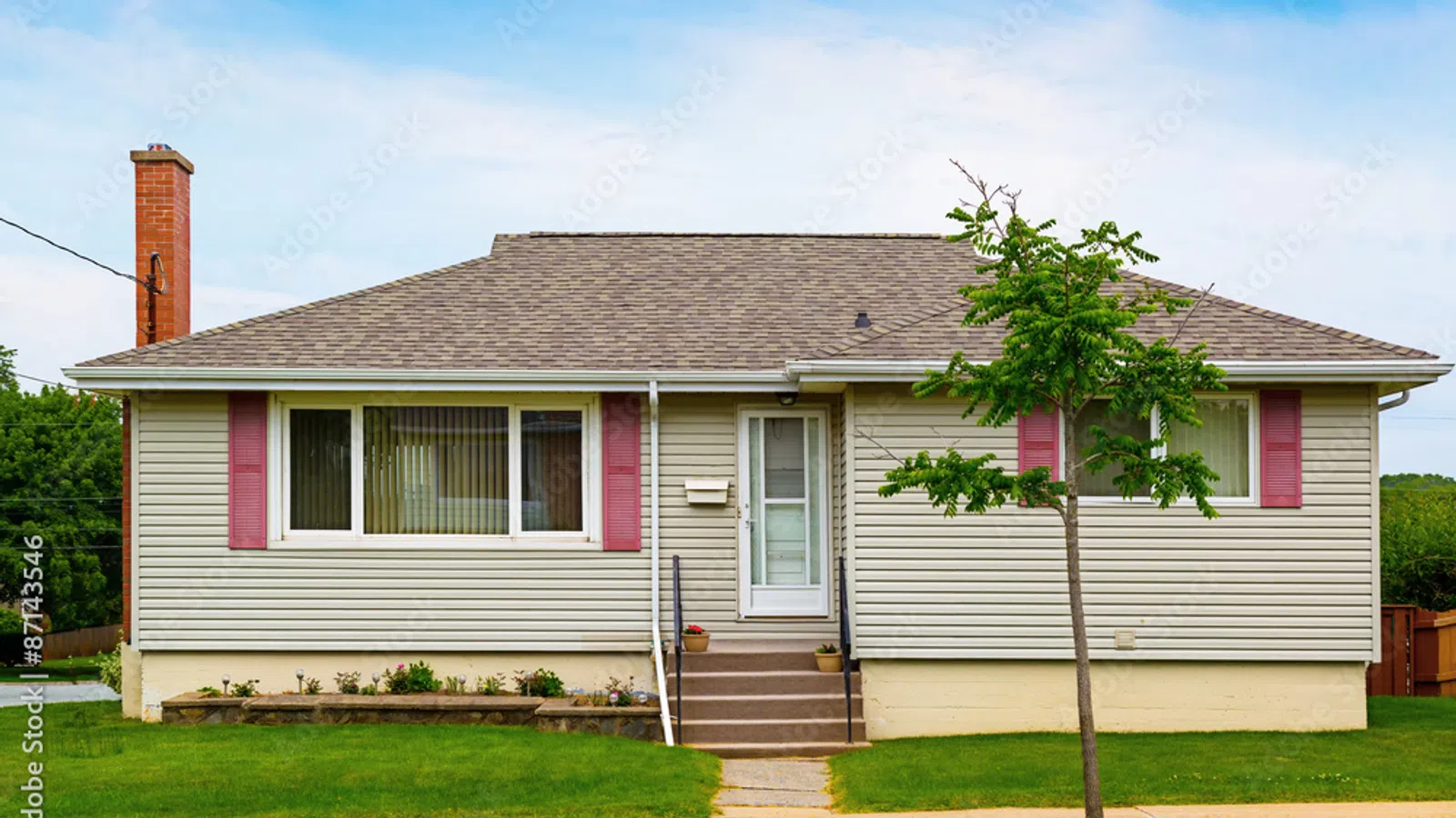 Single-story beige house with red shutters, chimney, front steps, small tree, and green lawn under blue sky.