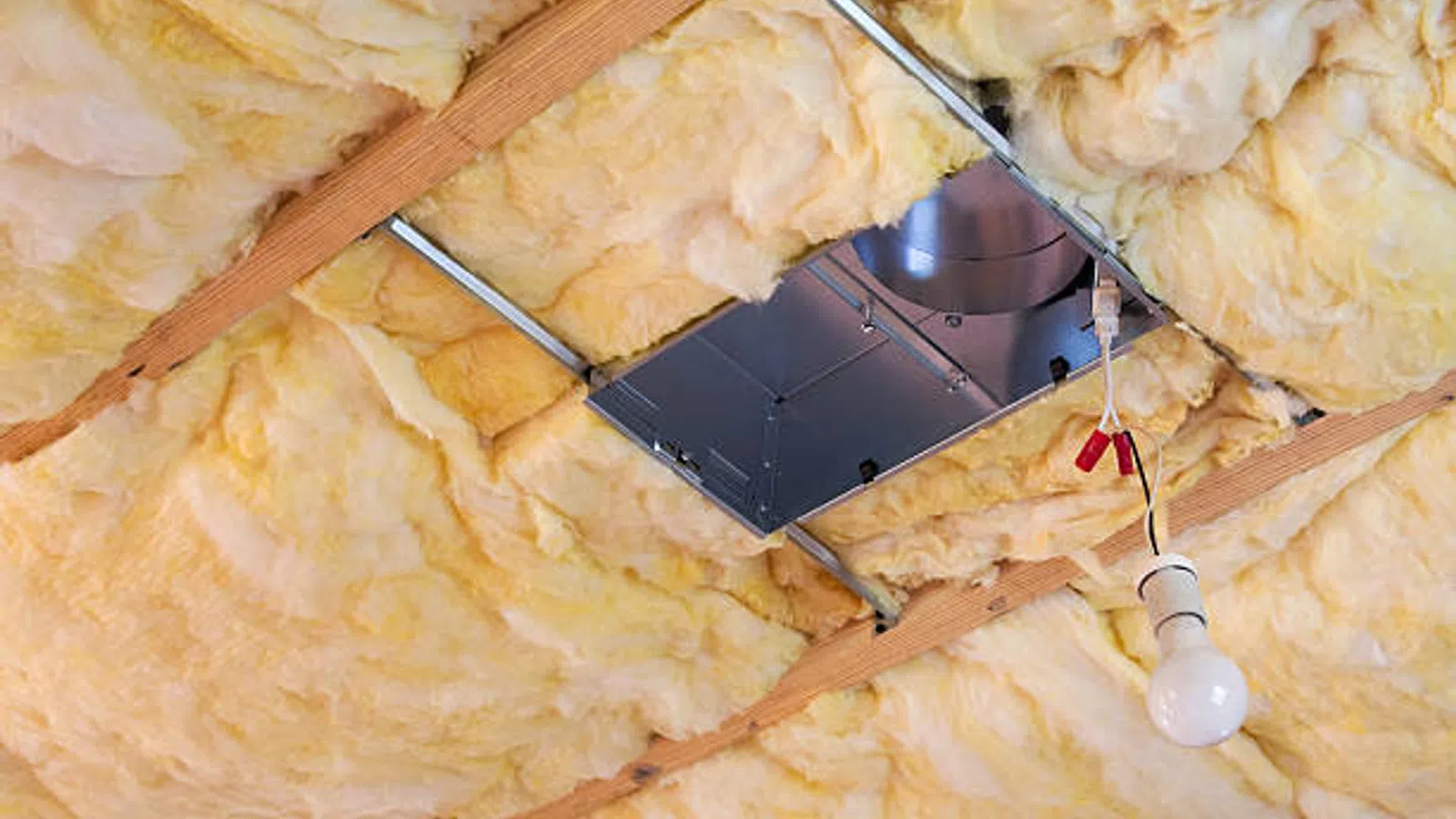 Yellow fiberglass insulation installed between wooden ceiling beams with a metal HVAC duct and hanging light bulb.