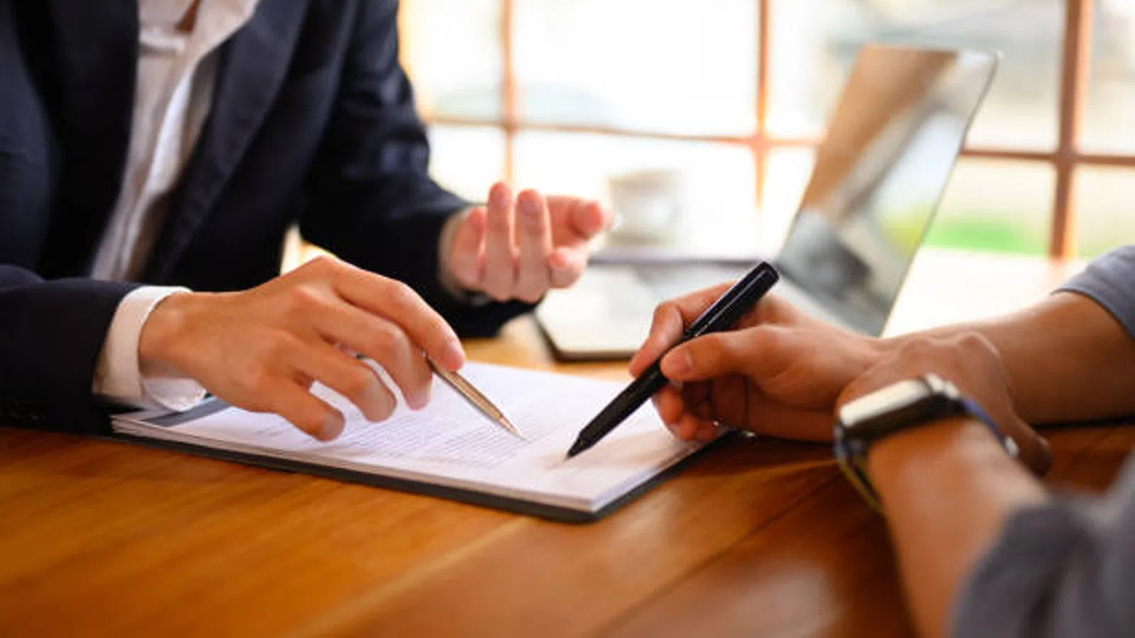 Two people reviewing and discussing a document with pens and a laptop on a wooden table.