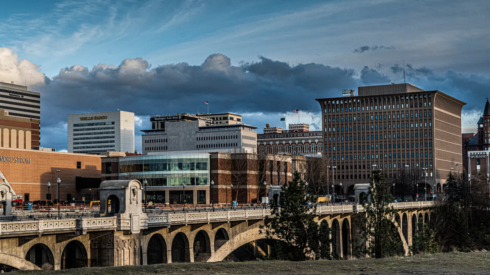 Downtown cityscape with historic bridge, modern buildings, and dramatic cloudy sky at sunset.