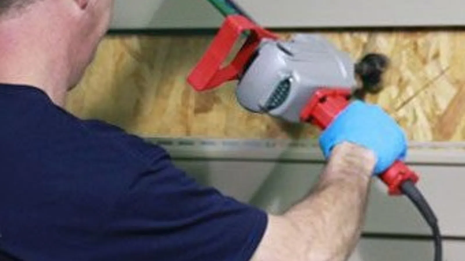 Man using a power tool to cut or remove siding from a wall with blue gloves and a wooden backing.