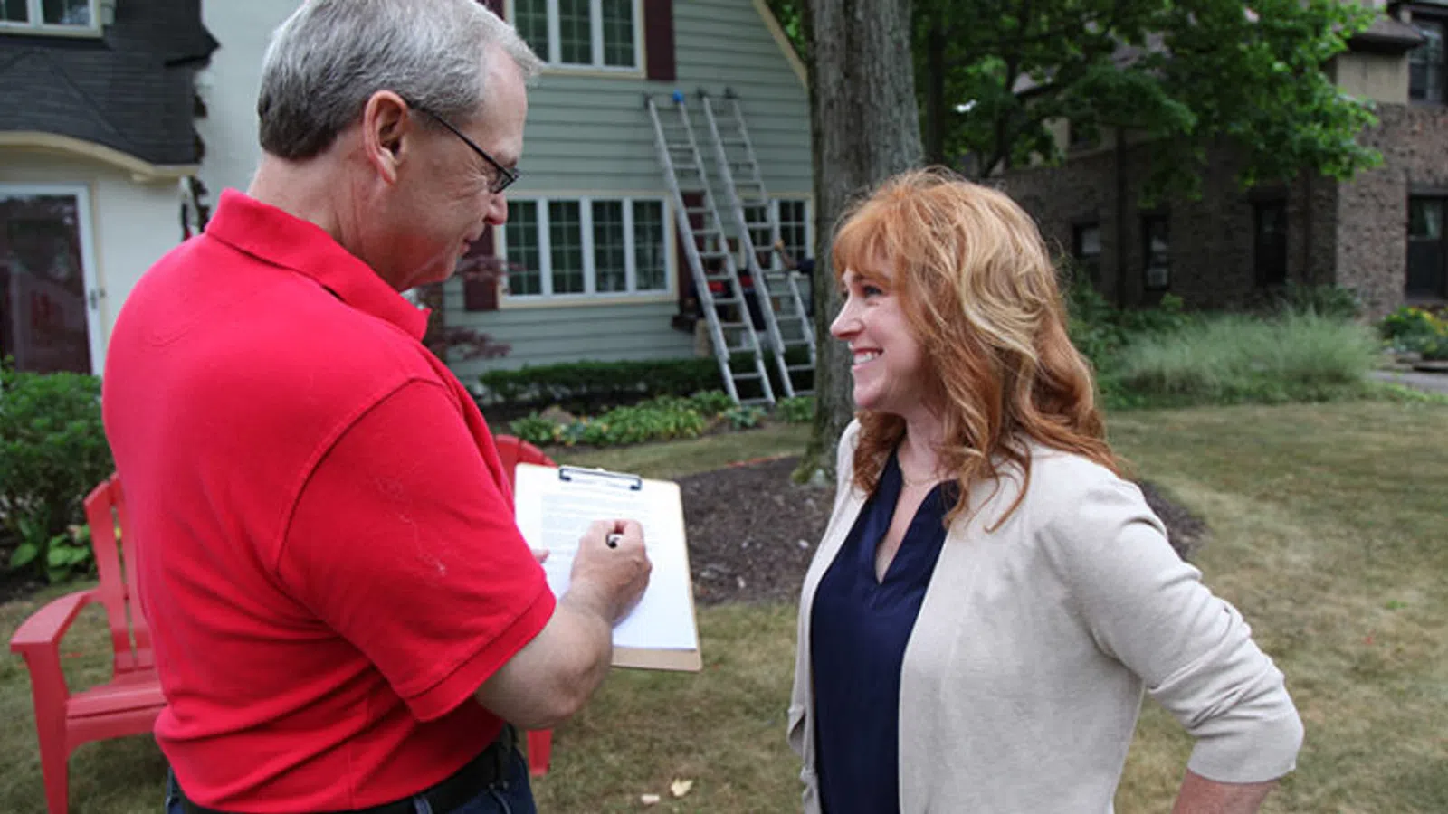 A man in a red shirt notes on a clipboard while talking to a woman outdoors in a residential area.