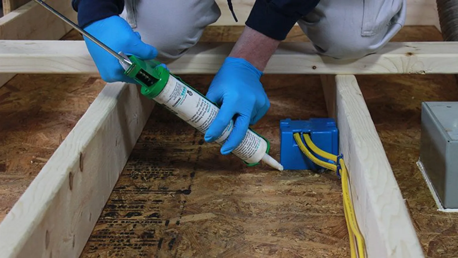 Person wearing blue gloves applying sealant around electrical box under wooden floor joists for safety.